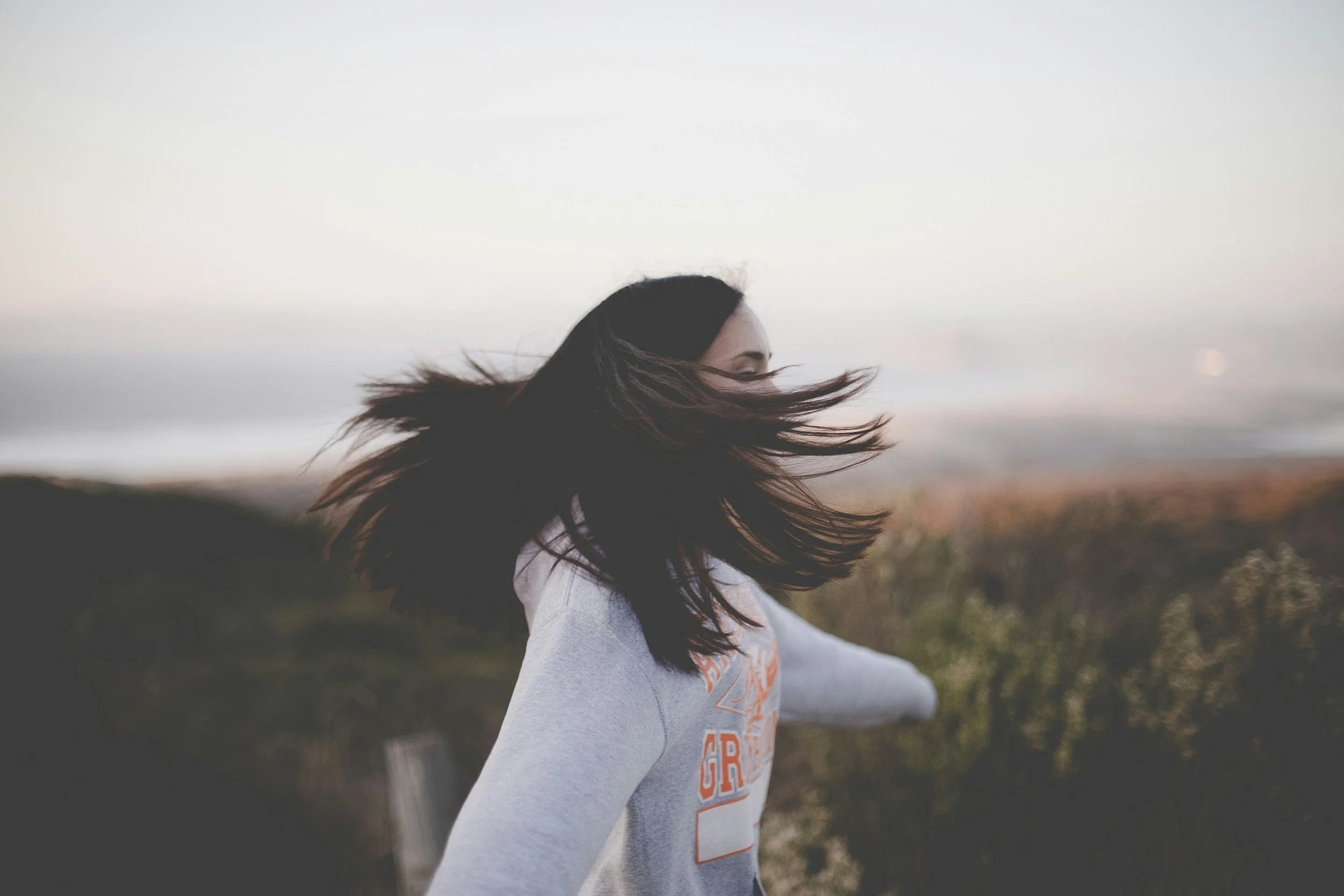 A woman with long dark hair blowing in the wind, standing outdoors with a cloudy sky in the background.
