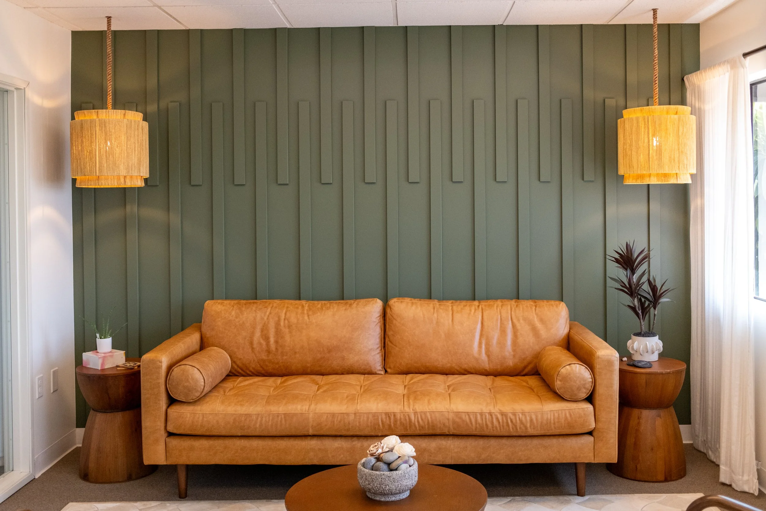 Living room with tan leather sofa, green textured wall behind it, two hanging pendant lights, side tables with plants, and a window with white curtains.