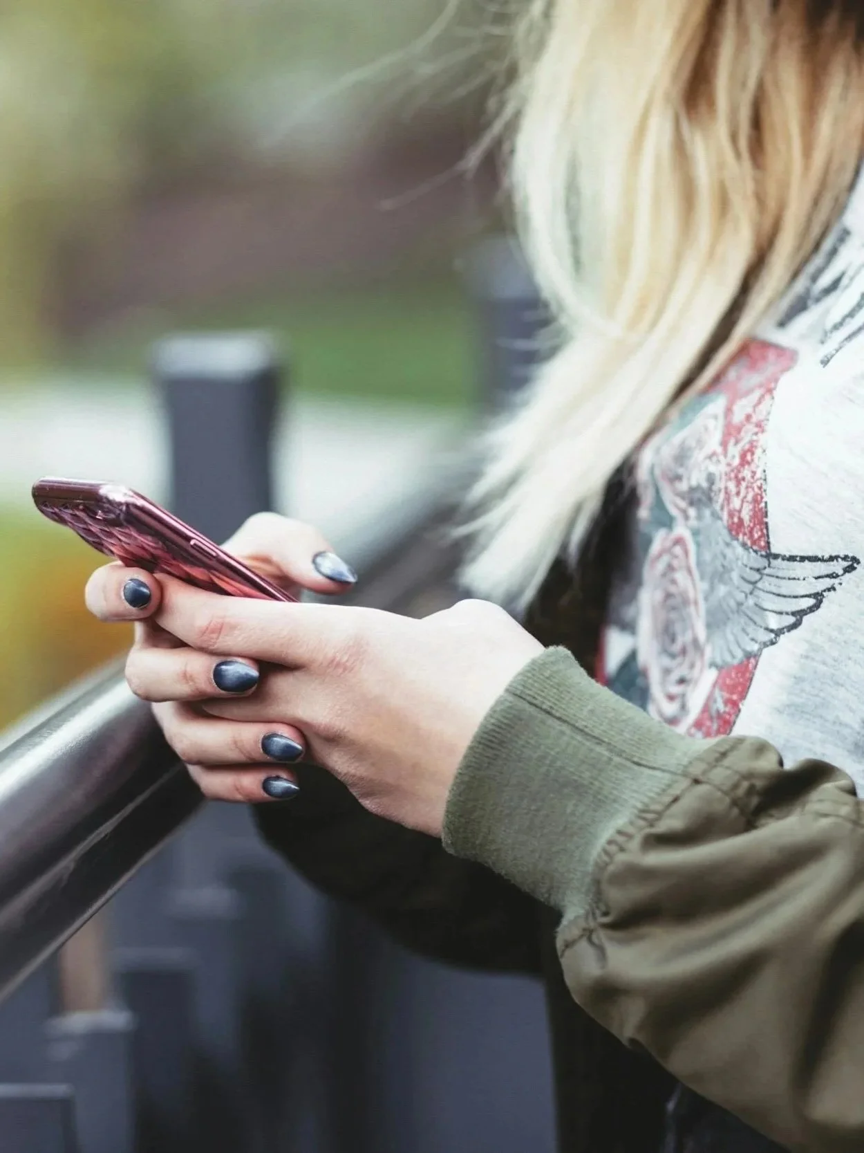 Close-up of a person holding and using a smartphone, wearing a green jacket and with dark painted nails, standing near a railing outdoors.