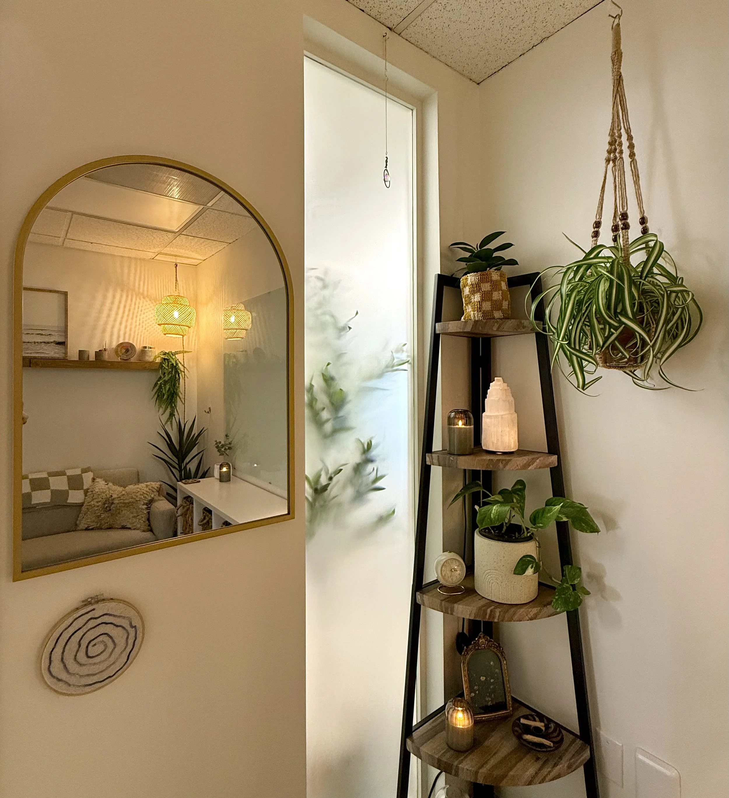 Interior of a cozy living space with a white wall, a tall ladder-style wooden shelf with potted plants and candles, a hanging potted plant, and a frosted glass door. The reflection in a gold-framed mirror shows a sofa with pillows, a side table, and hanging pendant lights.