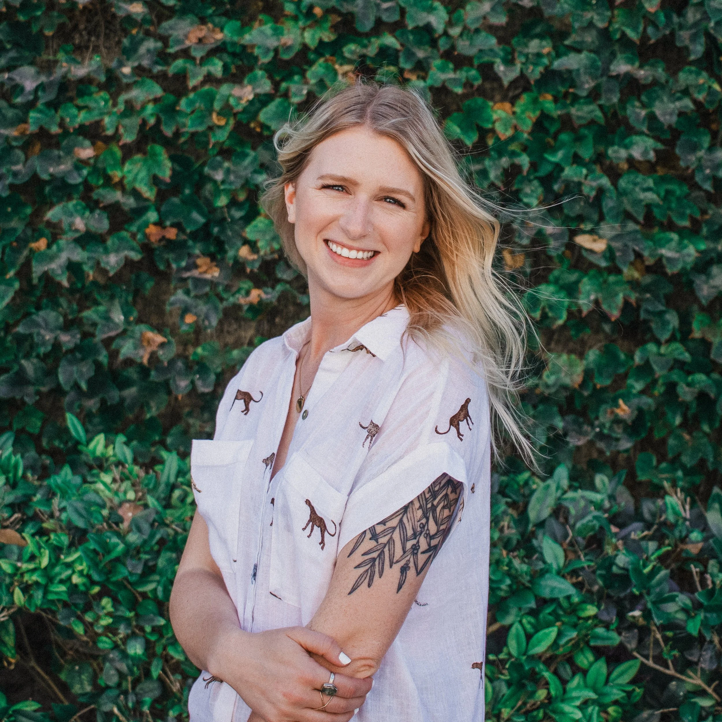 A smiling woman with blonde hair and a tattoo on her arm, standing outdoors in front of a leafy green background.