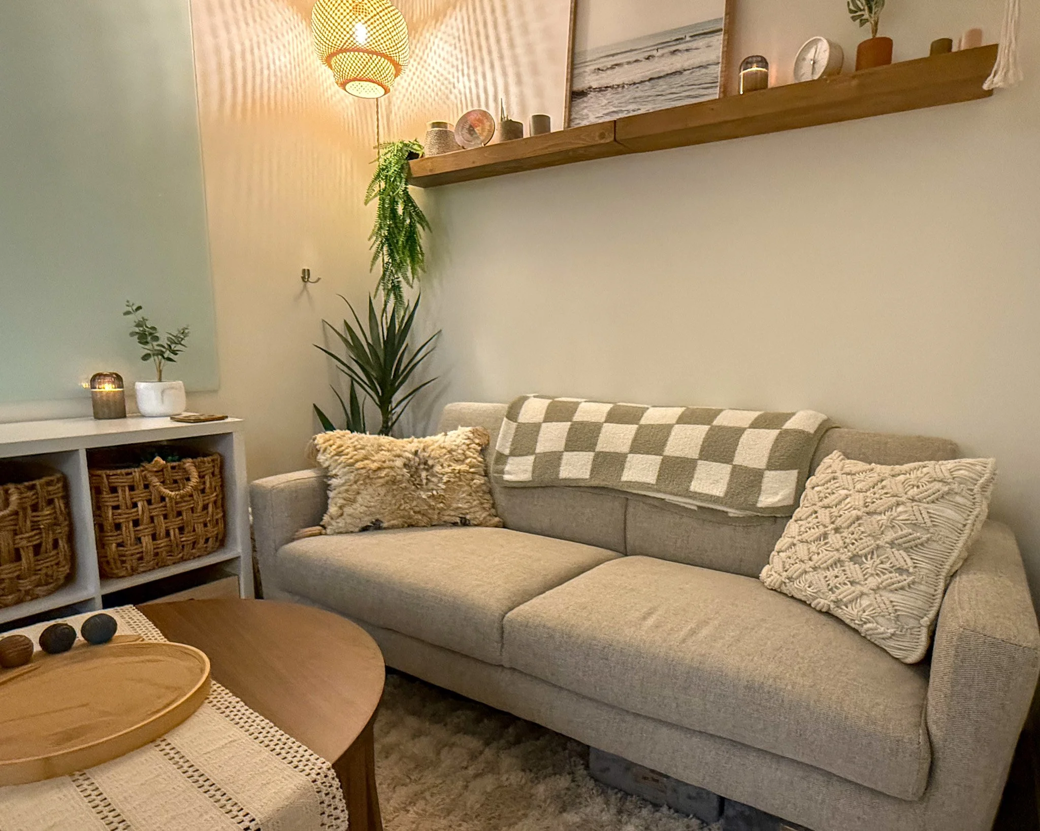 Cozy living room with a beige upholstered sofa decorated with textured pillows and a checkered throw blanket, a wooden coffee table with a decorative tray and balls, a white shelving unit with woven baskets, potted plants, a large green leafy plant, and wall art with candles and a floating wooden shelf with small decorative objects. Warm lighting from a pendant lamp adds to the inviting atmosphere.