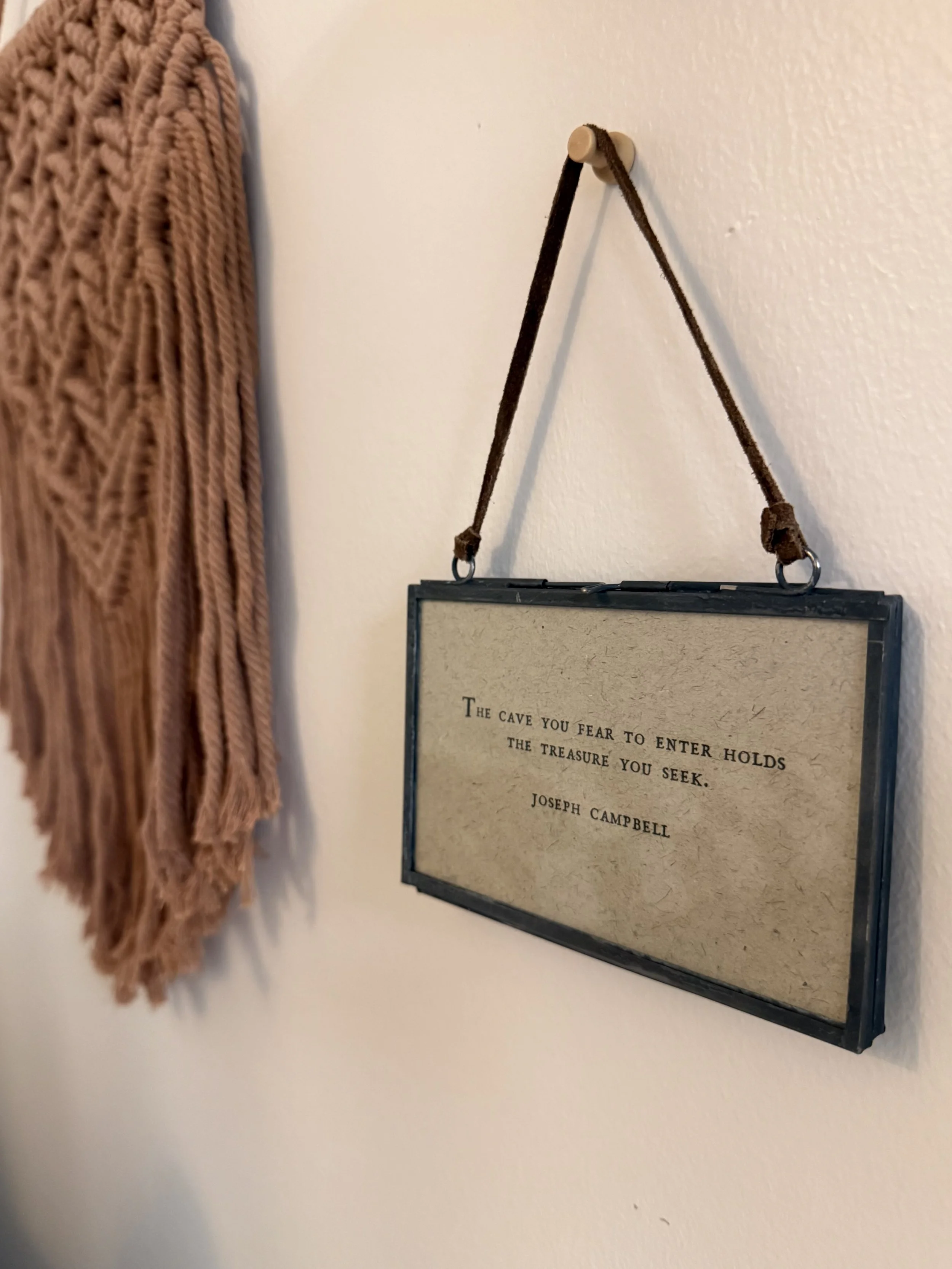 A framed quote hanging on a wall with a leather strap, next to a textured brown fabric hanging on the wall.