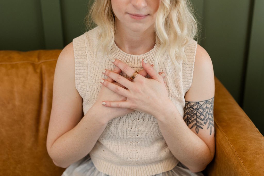 A woman with blonde, wavy hair and a floral tattoo on her right arm, sitting on a brown couch, wearing a sleeveless knitted beige top, holding her hands over her chest, with a gentle smile.