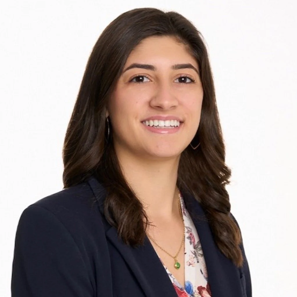 A young woman with shoulder-length brown hair, wearing a navy blazer and patterned blouse, smiling at the camera against a plain white background.