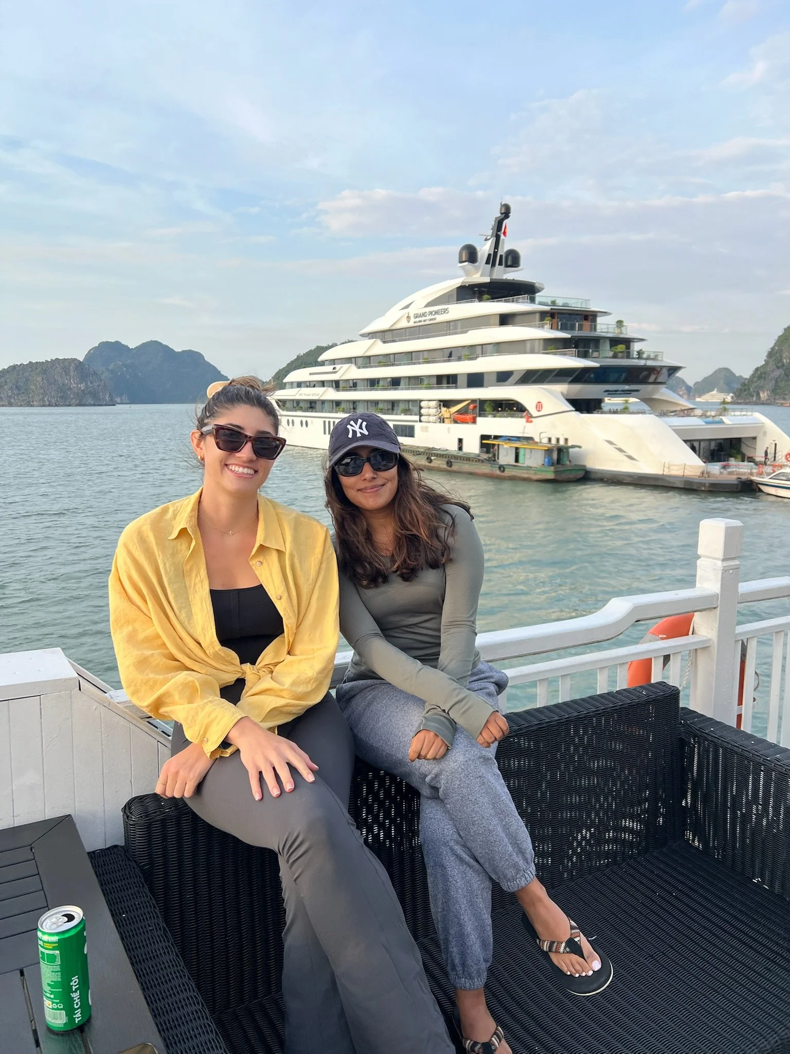 Two women sitting on a boat deck with a large yacht in the background, water, islands, and a partly cloudy sky.