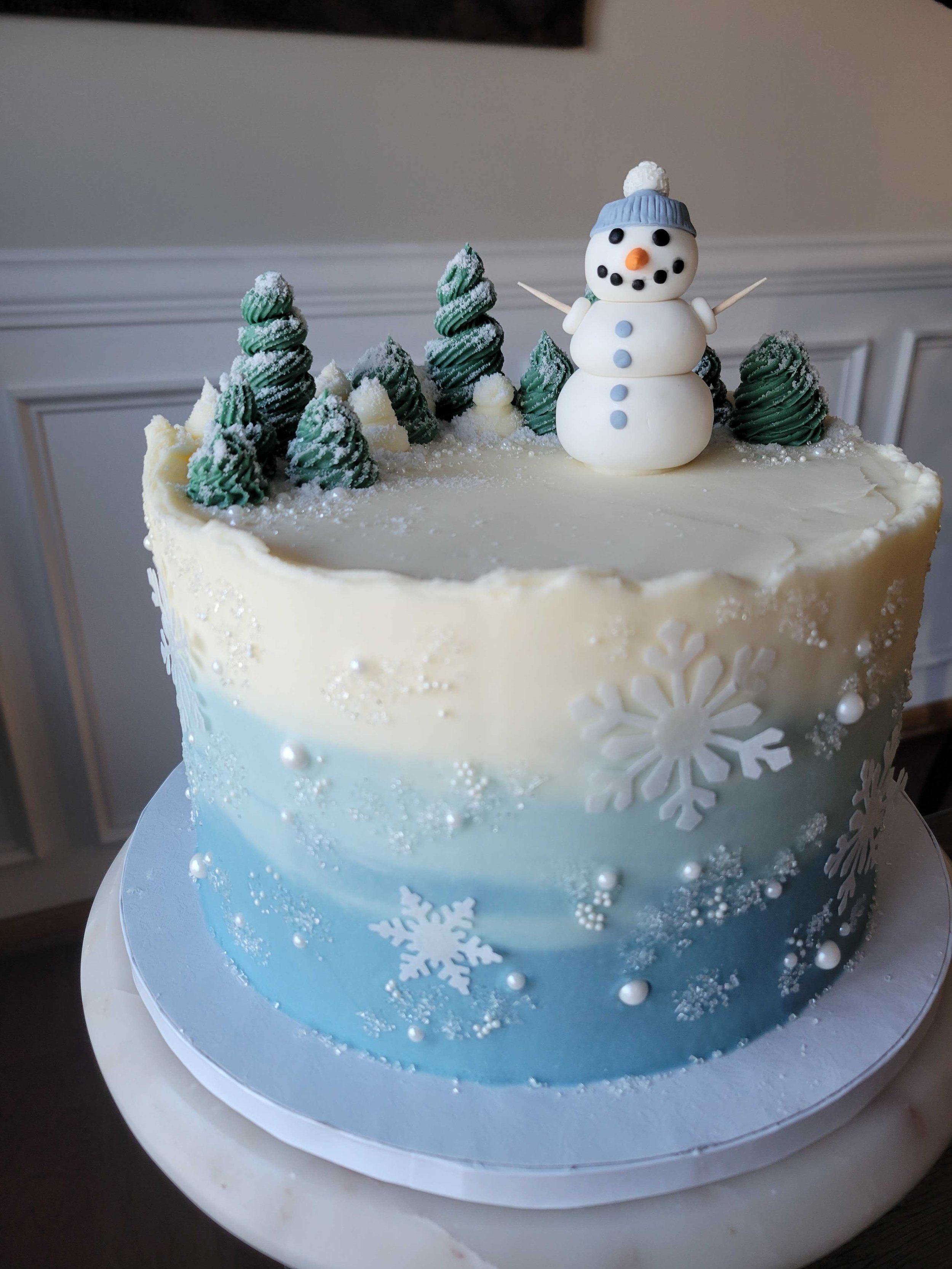 A snow-themed cake with white and blue icing, decorated with white snowflakes, small white pearls, and silver sugar beads. The top features a snowman made of white fondant with a blue hat, orange nose, and black eyes, with mini white fondant arms. There are small green frosted trees around the snowman on the cake.