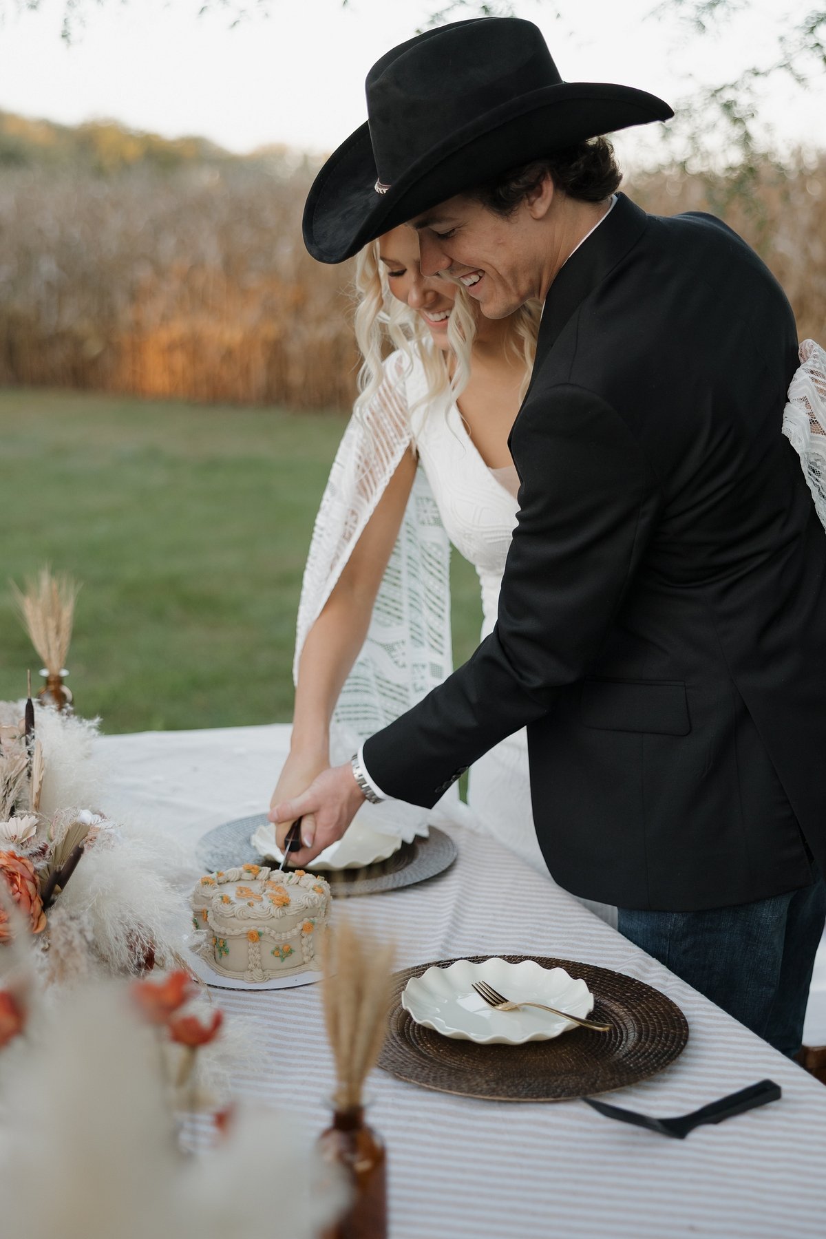 A couple cuts a wedding cake outdoors, both smiling and leaning toward each other, with a table decorated for a celebration.
