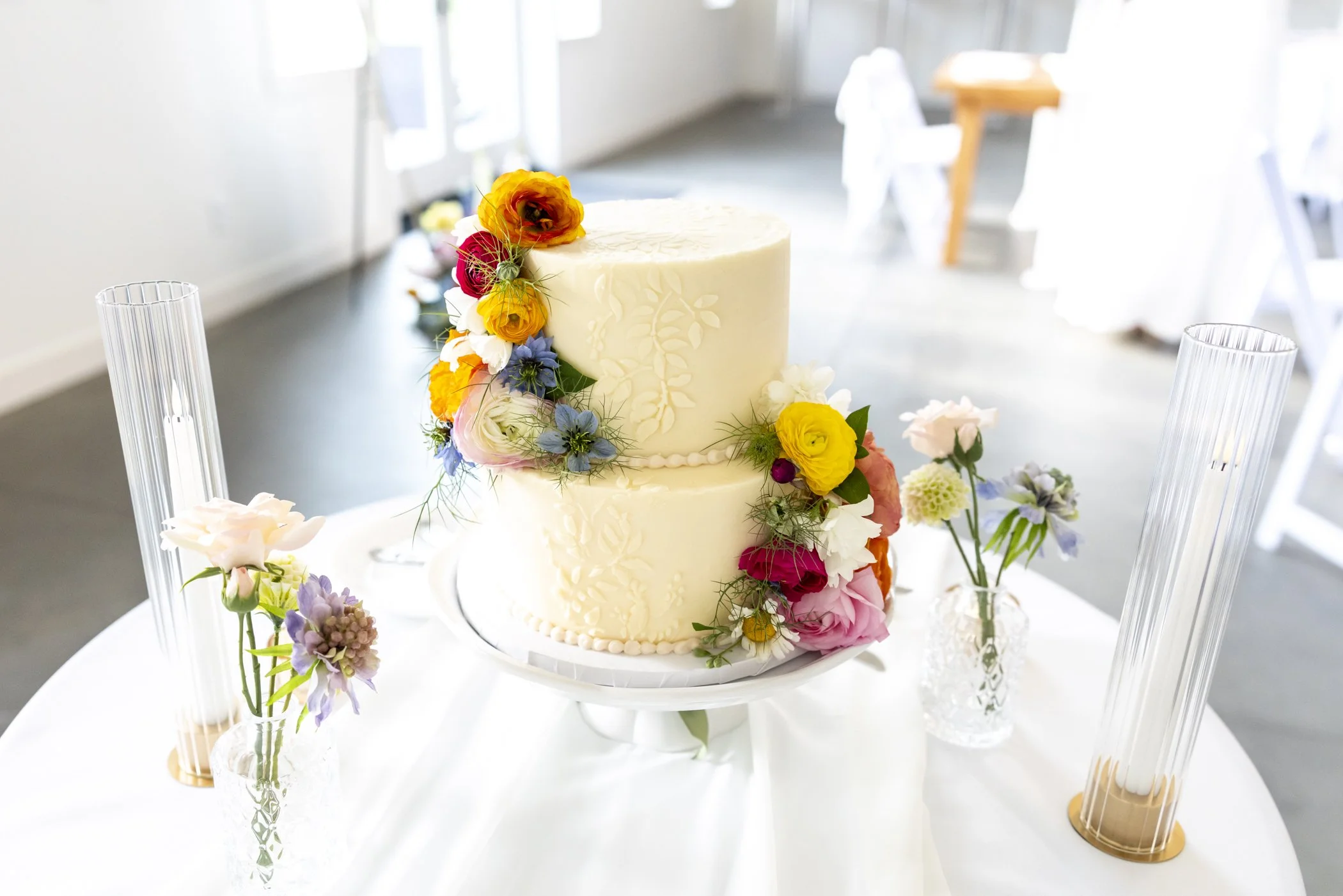 Two-tiered white wedding cake decorated with colorful fresh flowers, placed on a white table with glass vases and candles in a bright indoor setting.