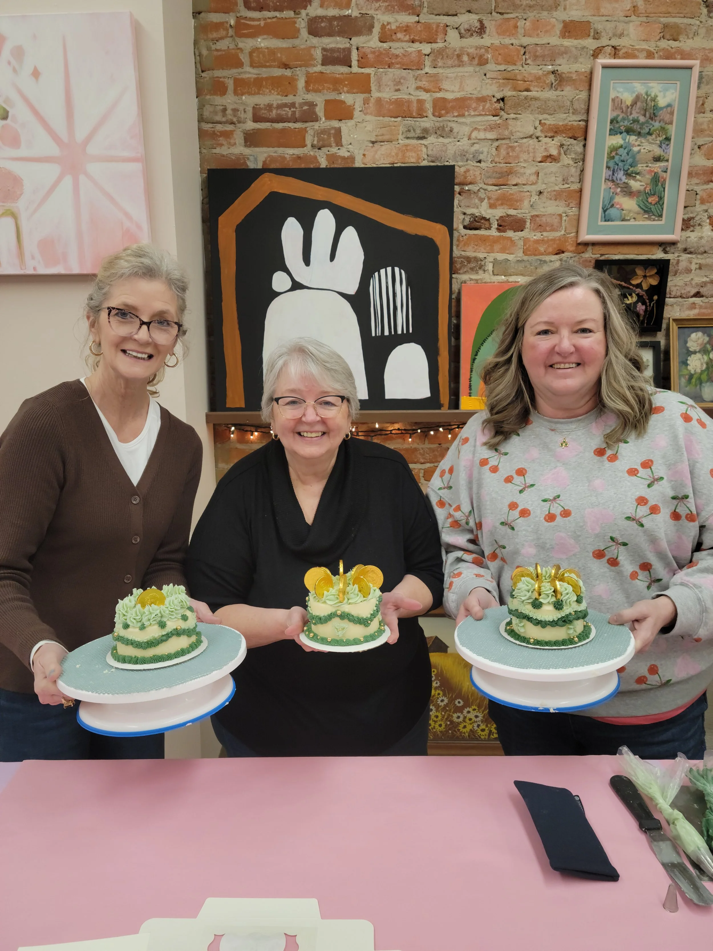 Three women smiling and holding decorated cakes, standing behind a pink table with a knife and napkin, in a room with artwork on the brick wall.