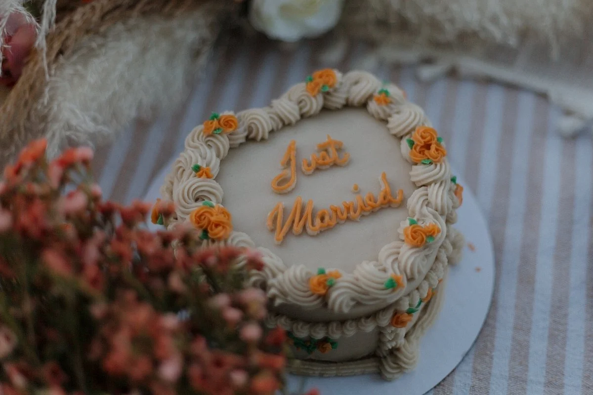A round wedding cake with white frosting, decorated with orange and green flower icing, with the words "Just Married" written in orange icing on top, sitting on a striped fabric tablecloth.