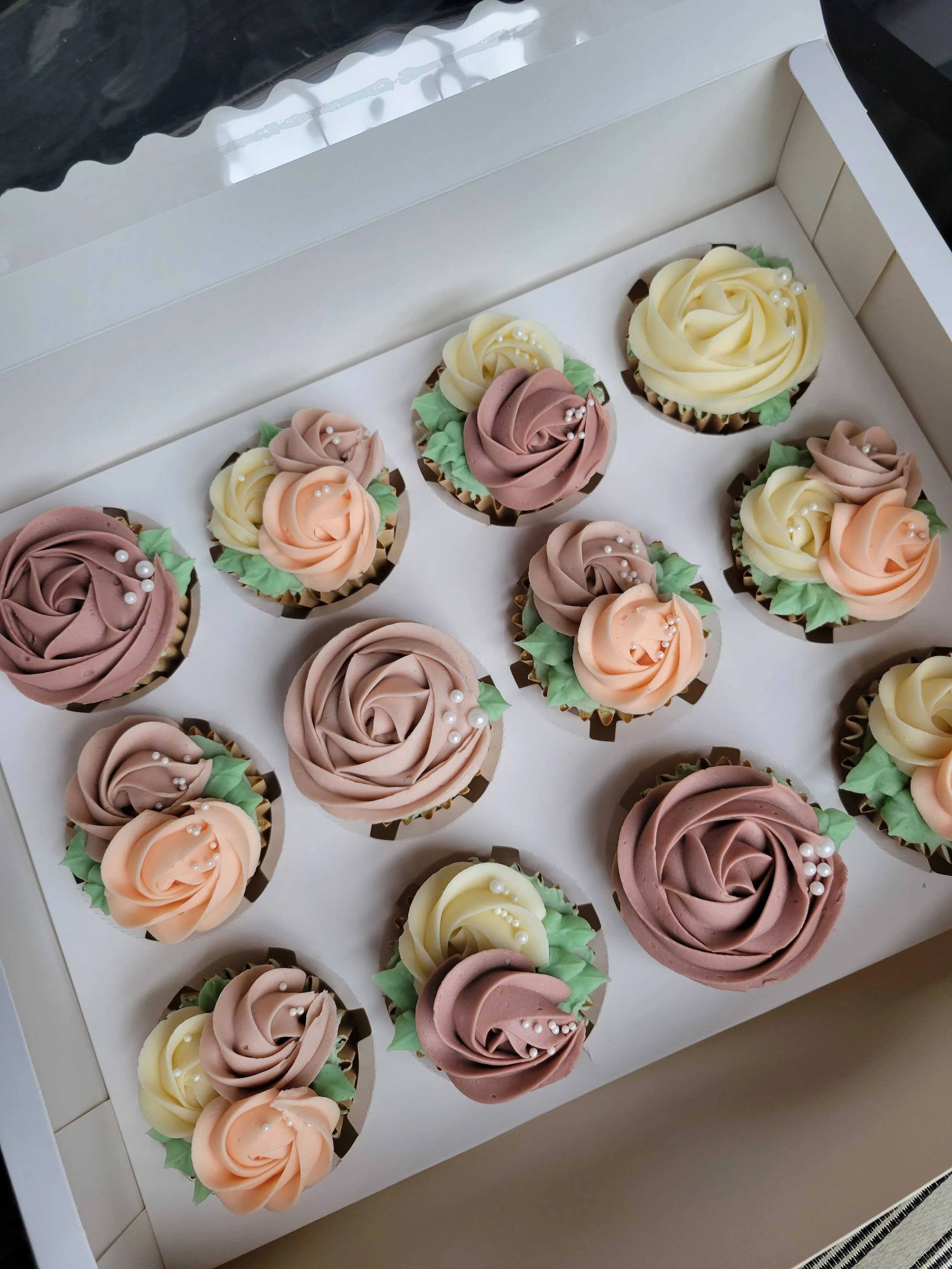 A white box containing an assortment of cupcakes decorated with rose-shaped frosting in shades of brown, cream, peach, and yellow, decorated with white edible pearls and green leaves.