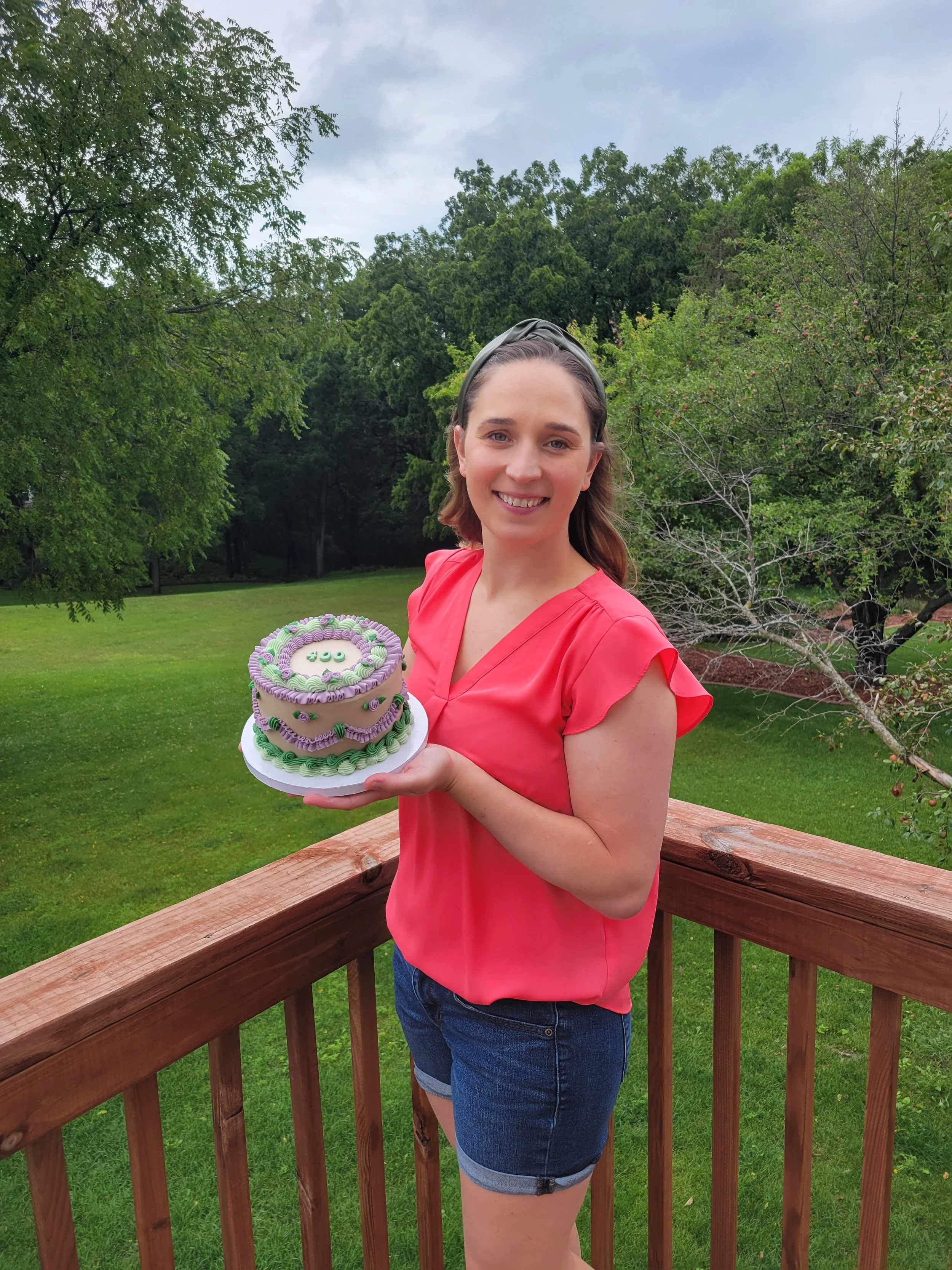 A young woman with light skin and brown hair smiling, holding a decorated cake, standing outdoors on a wooden deck with green grass and trees in the background.