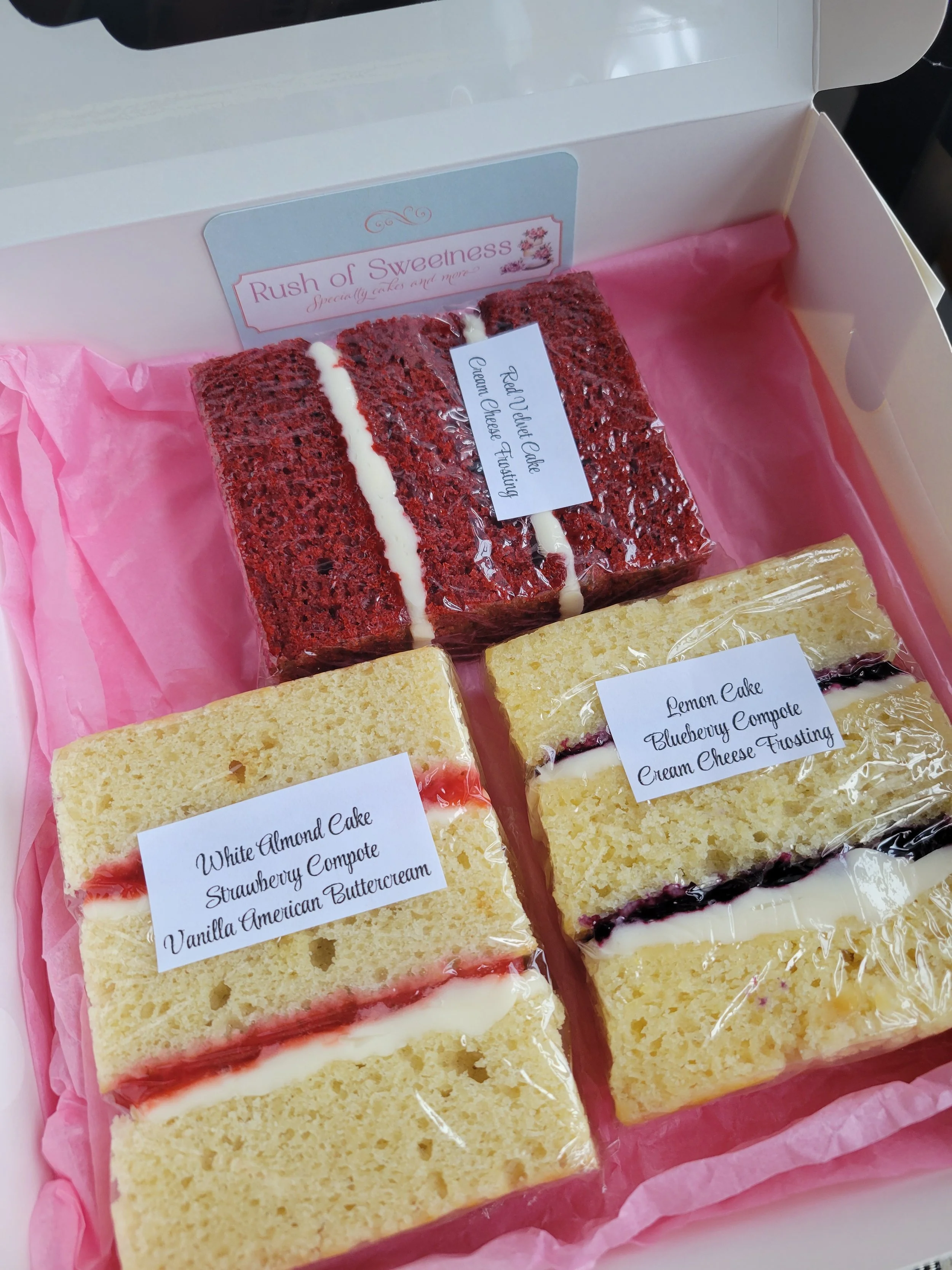 Assorted slices of cake in a box, including Red velvet cake, white almond cake, and lemon cake with blueberry compote and cream cheese frosting.