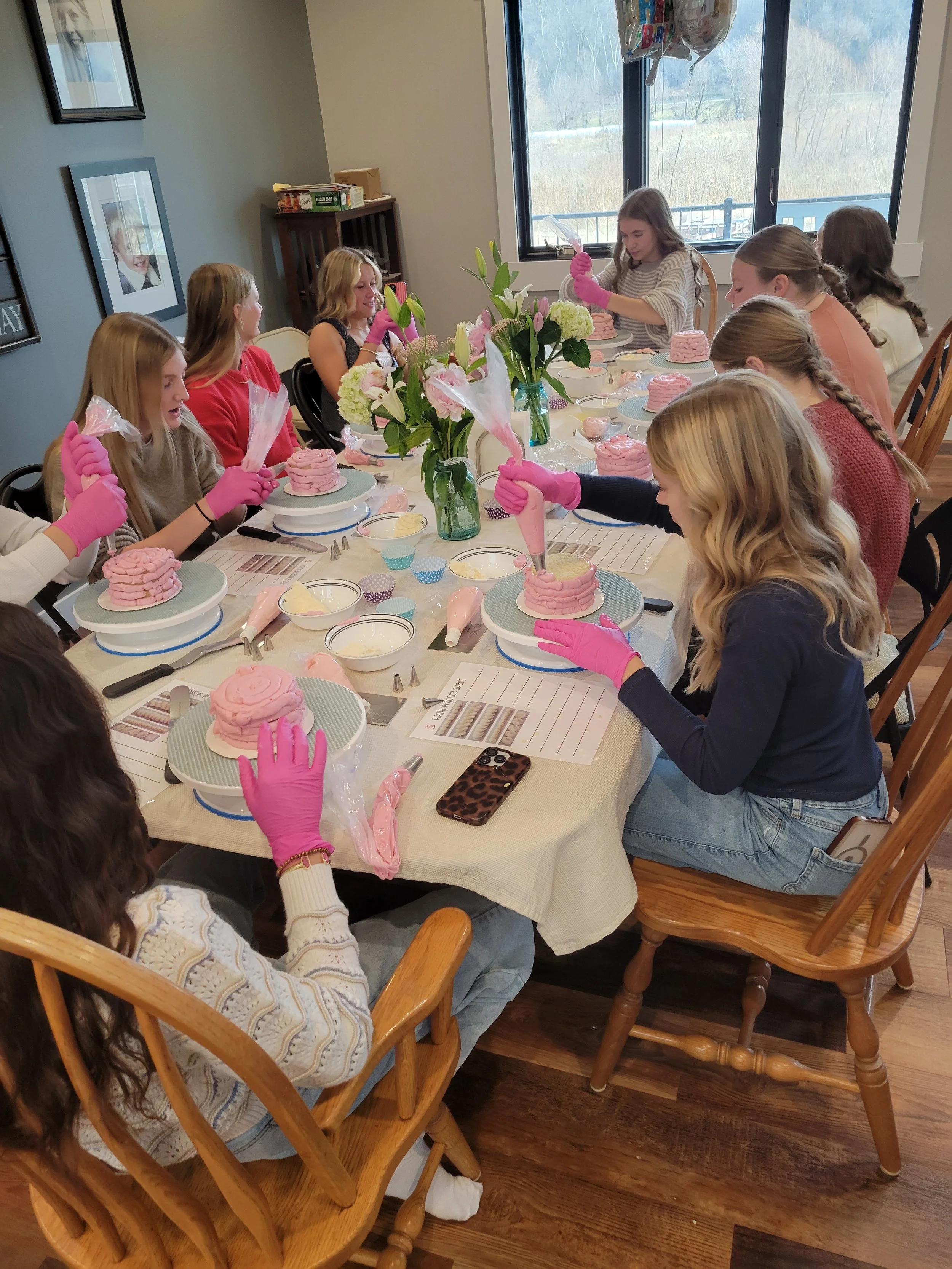 A group of girls at a birthday party decorating pink-frosted cakes, wearing pink gloves, seated around a dining table with flowers in vases and party balloons in the background.