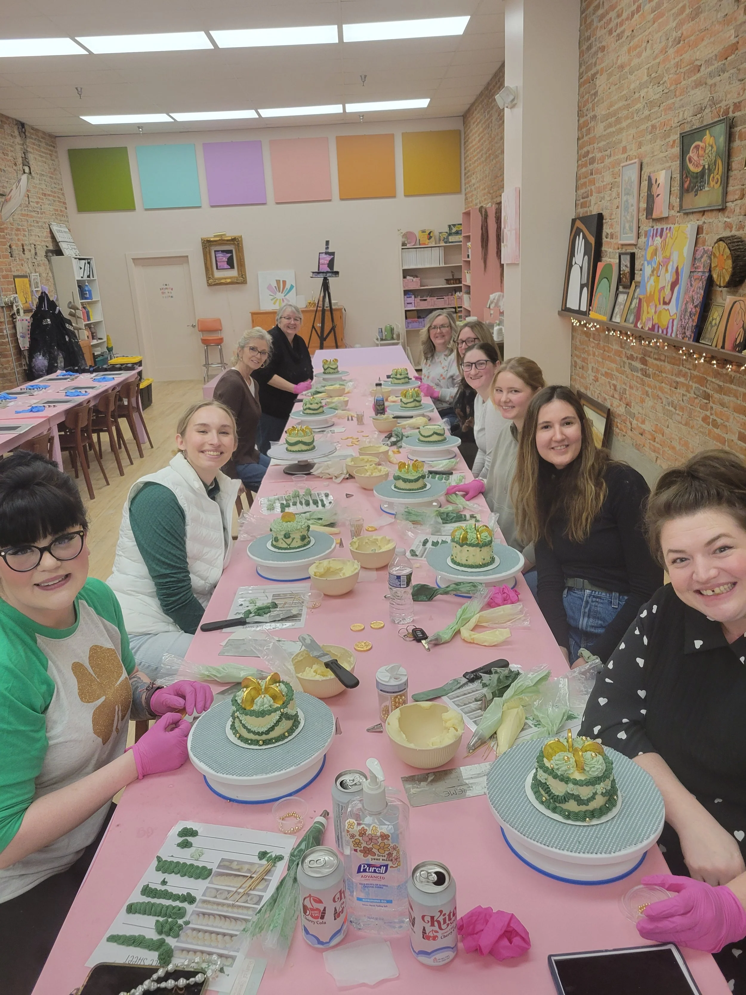Group of women sitting around a pink table decorating cakes with green and yellow icing in a decorated art studio.