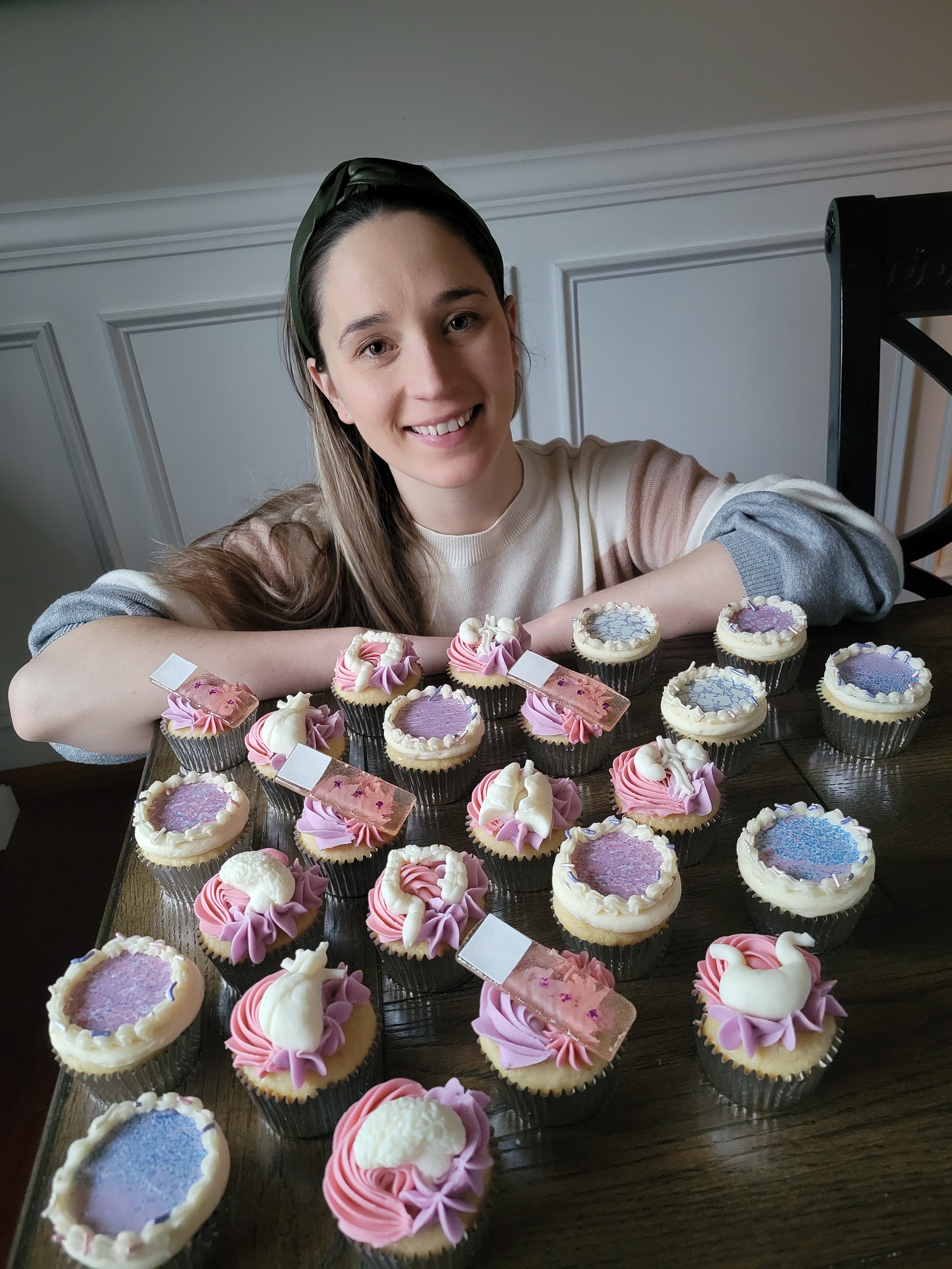A woman with long brown hair and a green headband smiling, sitting behind a table with pastel-colored cupcakes decorated with pink, purple, and white frosting, some with swan-shaped fondant toppers.