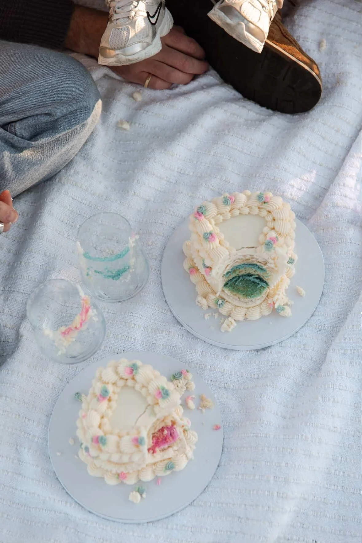 Two partially eaten layered birthday cakes with white frosting and pink and blue decorations on a white tablecloth, with empty glass cups nearby, and a person wearing jeans and sneakers sitting on the table.