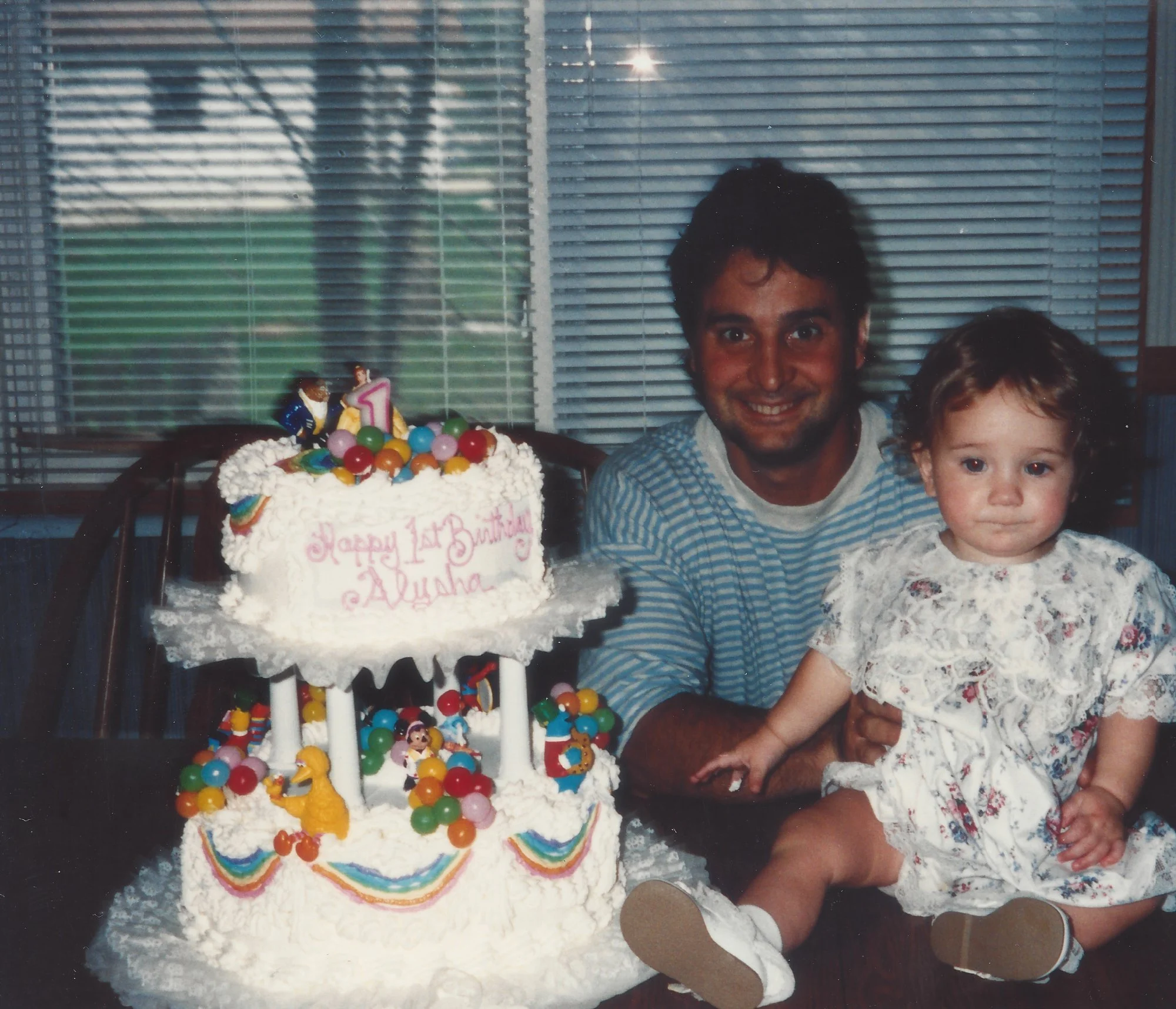 A man and a young girl sitting at a table with a large decorated birthday cake, which has a birthday message and figures, on a table. The man is smiling and the girl looks at the camera.