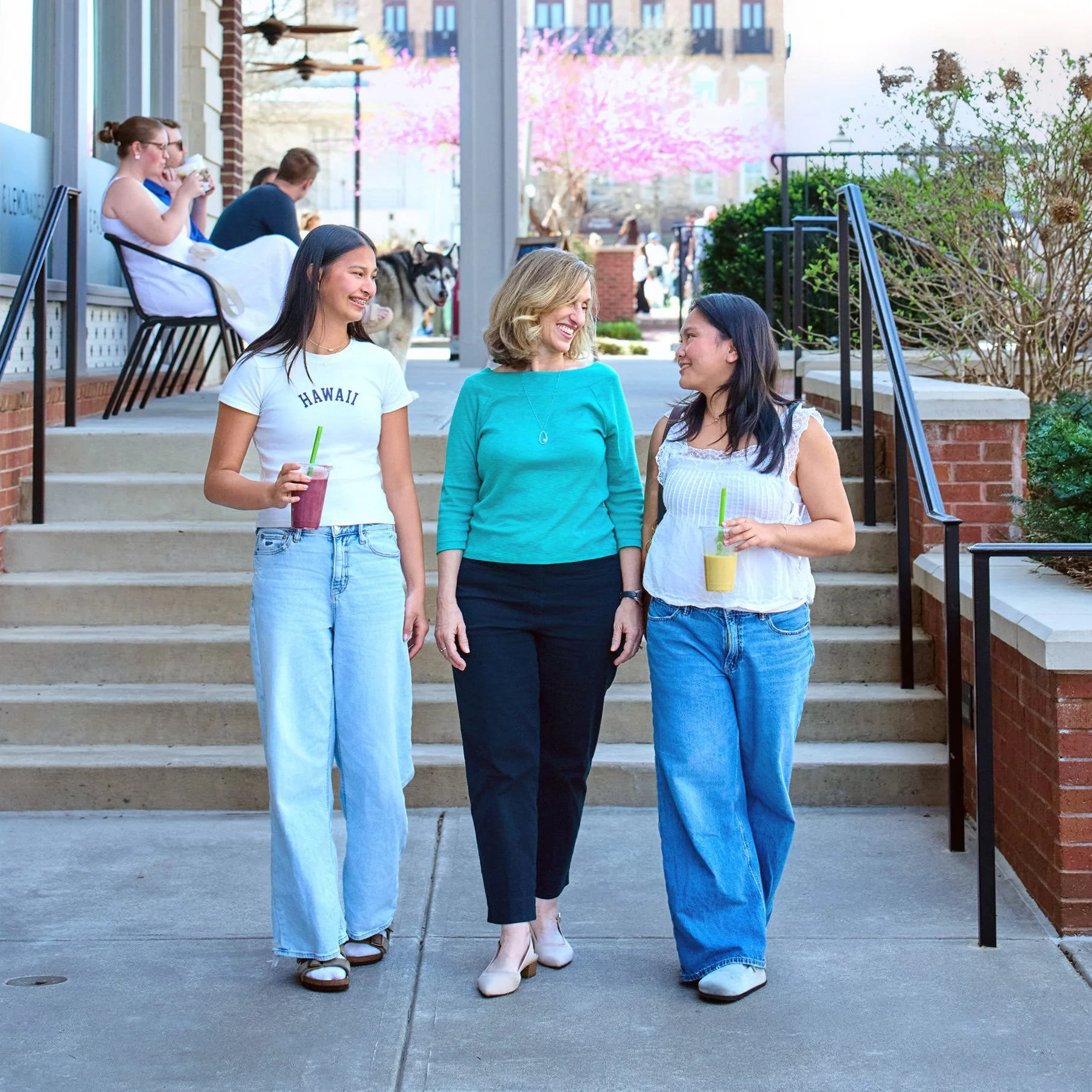 Three women walking down steps outside of a building, engaging in conversation and smiling, with others sitting on a bench in the background.
