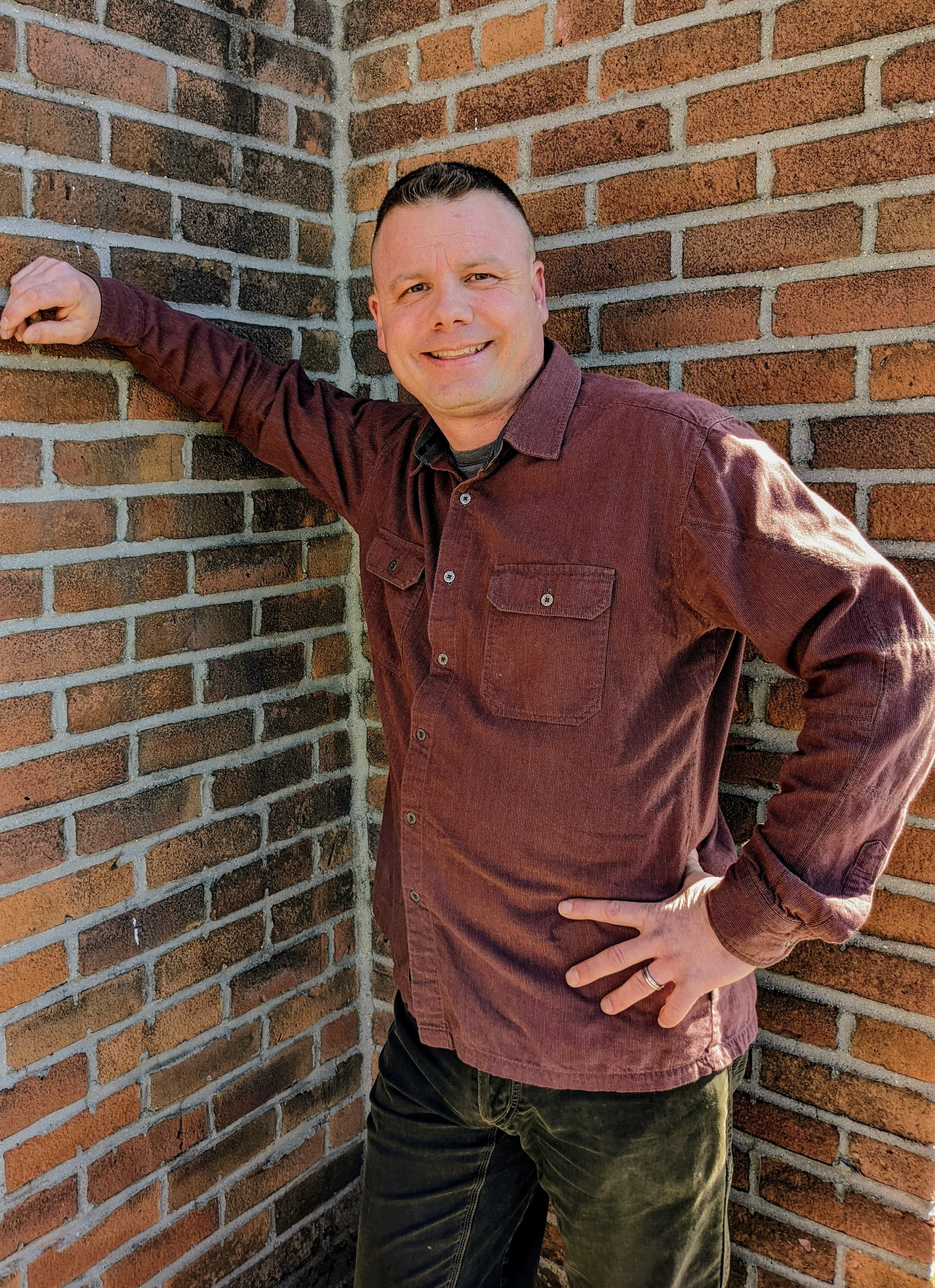 A man in a maroon shirt standing against a corner brick wall, smiling and looking at the camera.