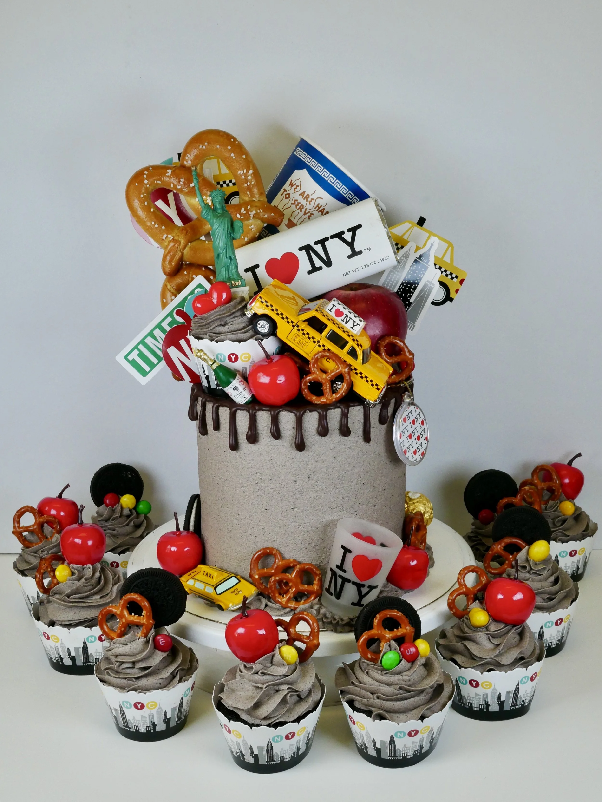 A cake decorated with New York City-themed items, including a miniature Statue of Liberty, taxis, and 'I ❤️ NY' signs, surrounded by chocolate cupcakes topped with cherries, pretzels, Oreos, and colorful candies.