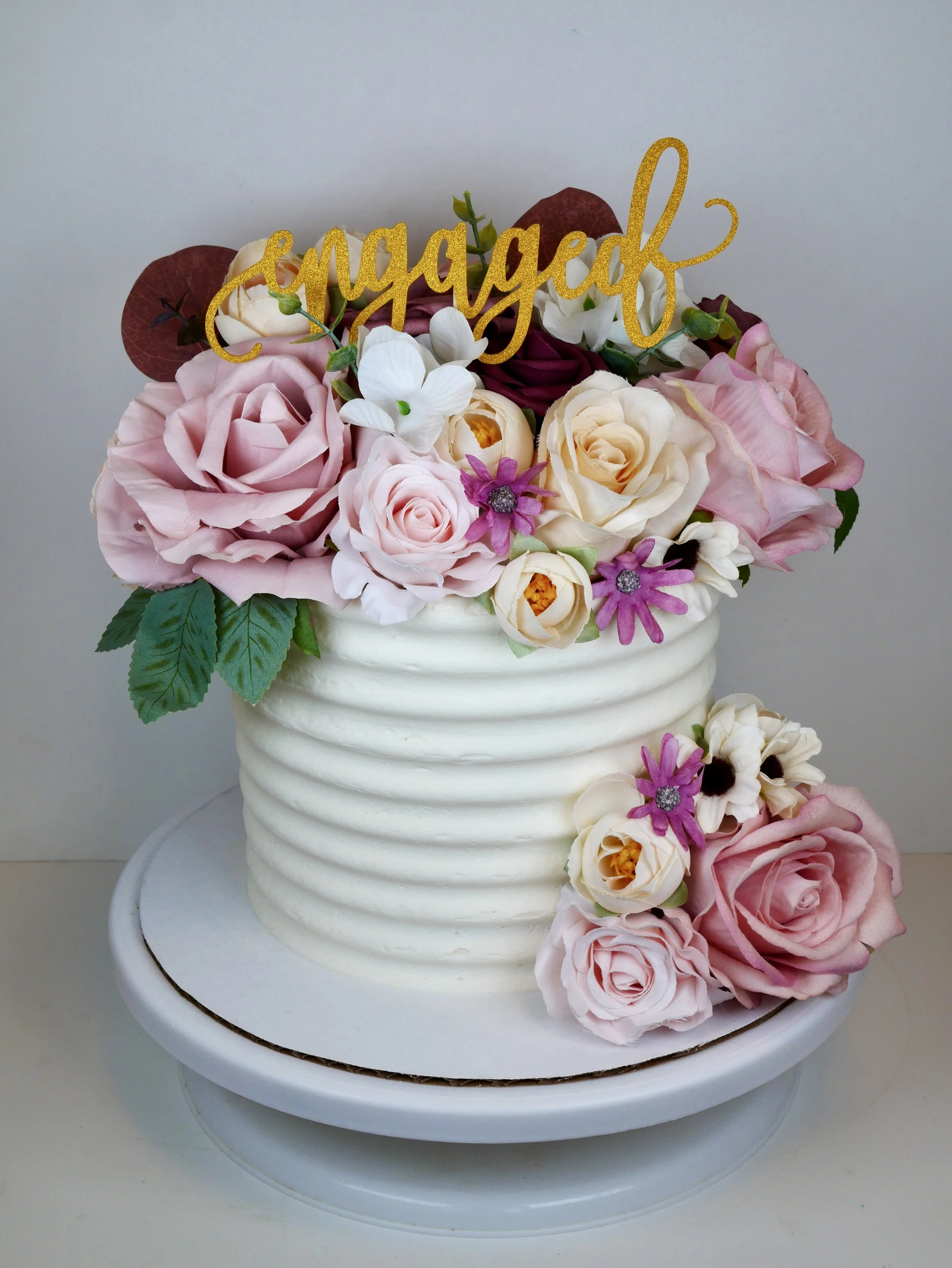A wedding cake decorated with pink and white roses, purple daisies, and white flowers, topped with a gold 'engaged' cake topper.