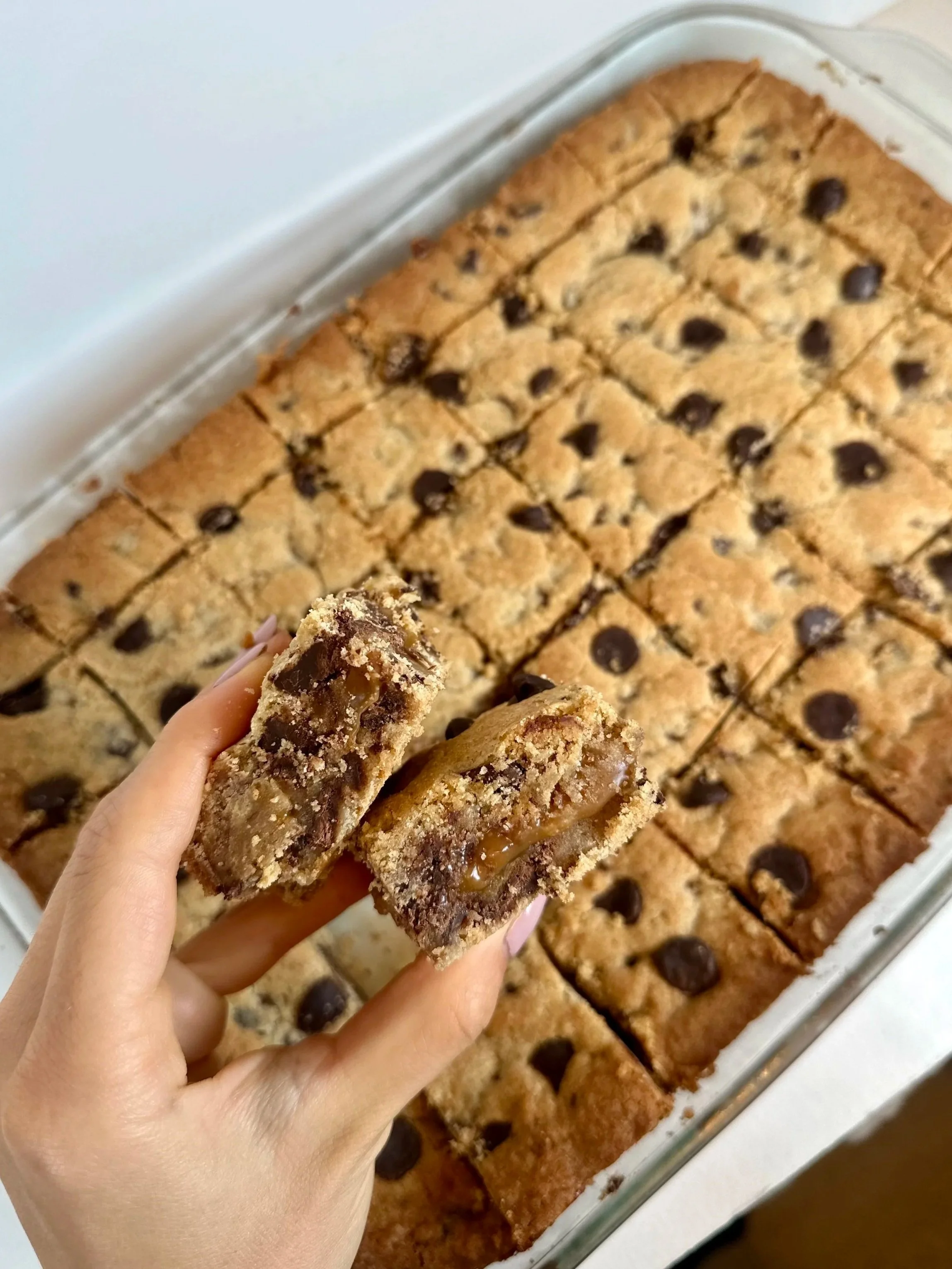 Close-up of a hand holding two pieces of cookie bar with caramel and chocolate chips, in front of a large tray of similar cookie bars.