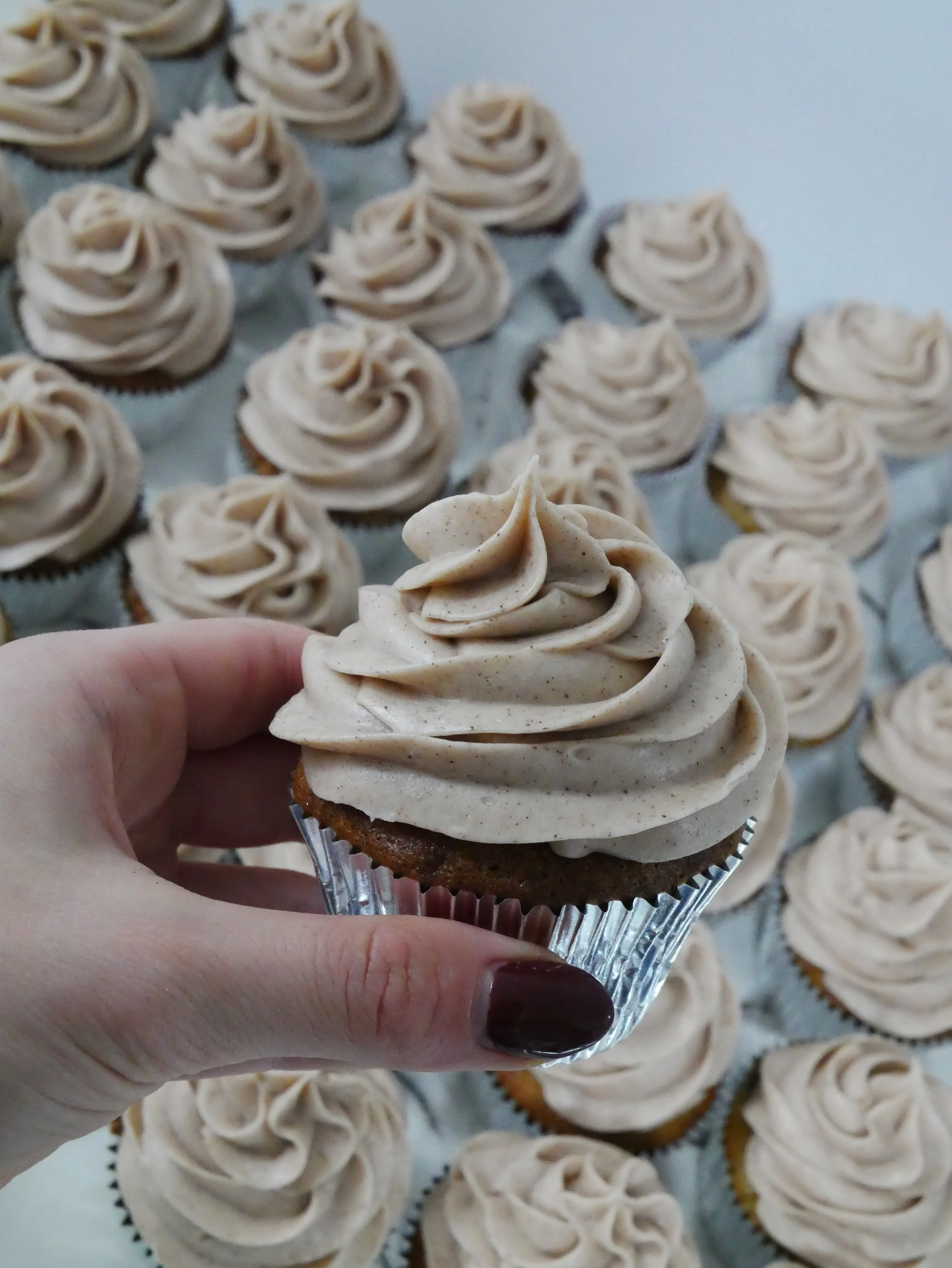 Hand holding a cupcake with beige frosting in front of a tray of similar cupcakes.