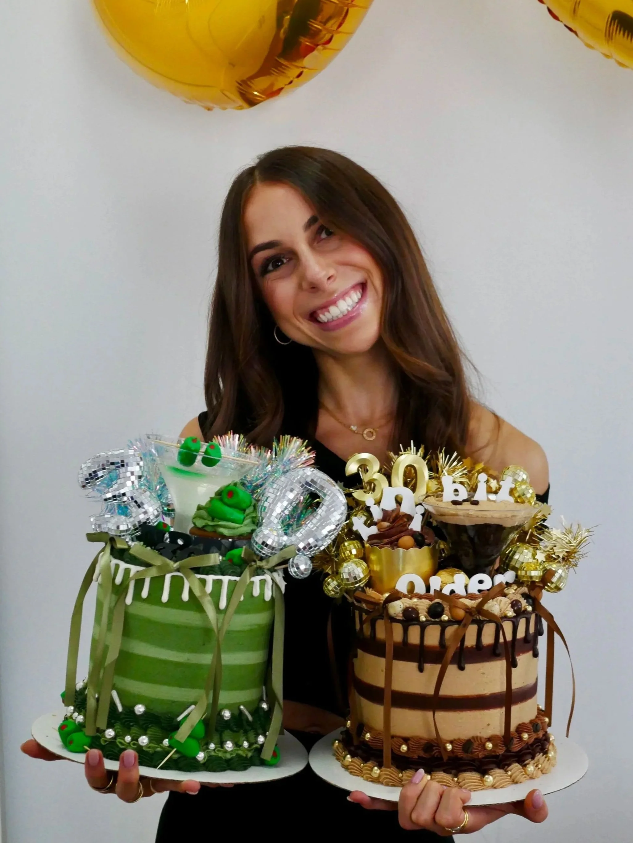 A woman celebrating her 30th birthday, holding two decorated birthday cakes with candles and decorations, standing in front of a white wall with partial gold balloons visible at the top.