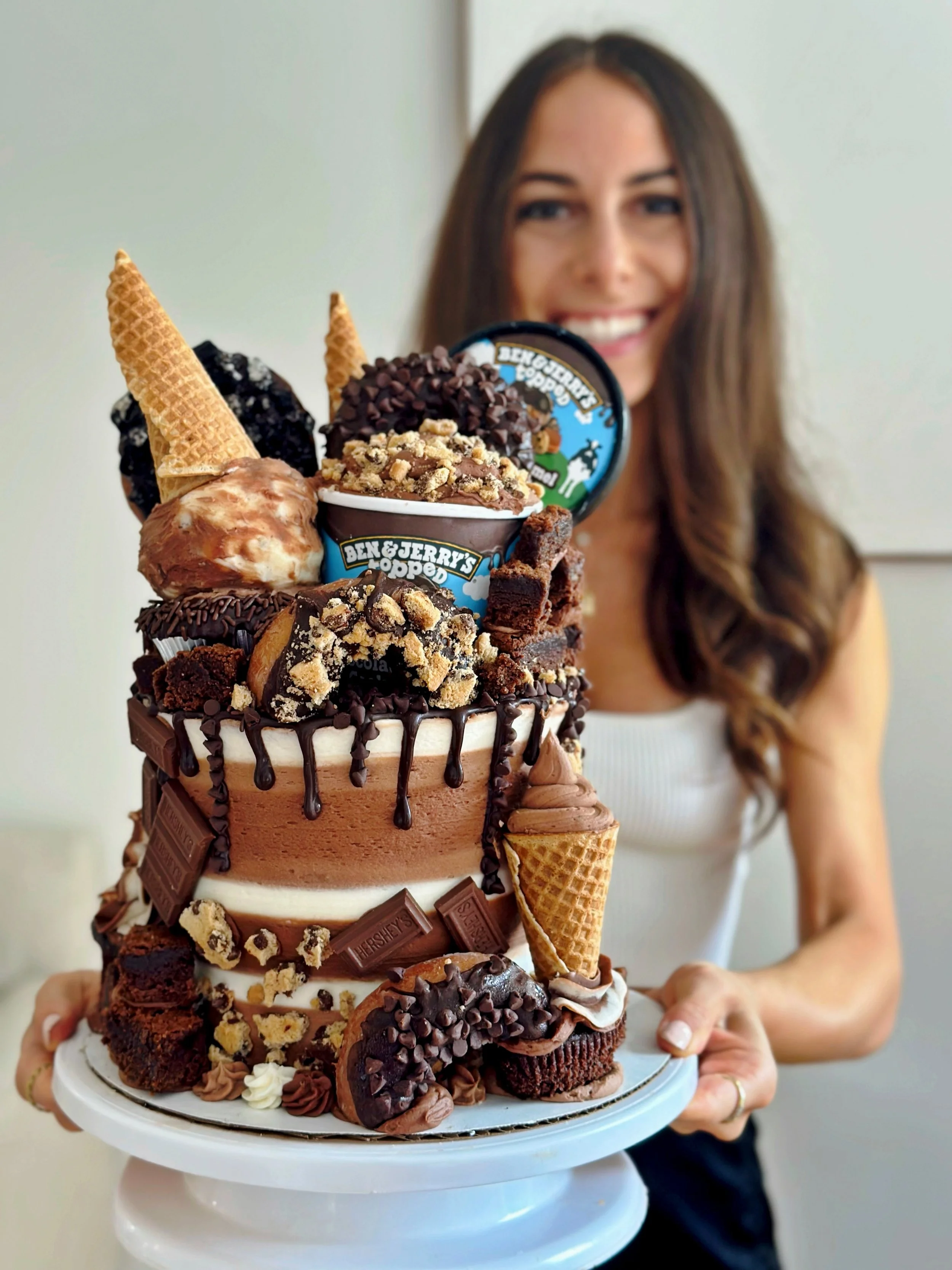 A woman holding a large, multi-layered ice cream cake decorated with chocolate, cookies, and ice cream tubs.