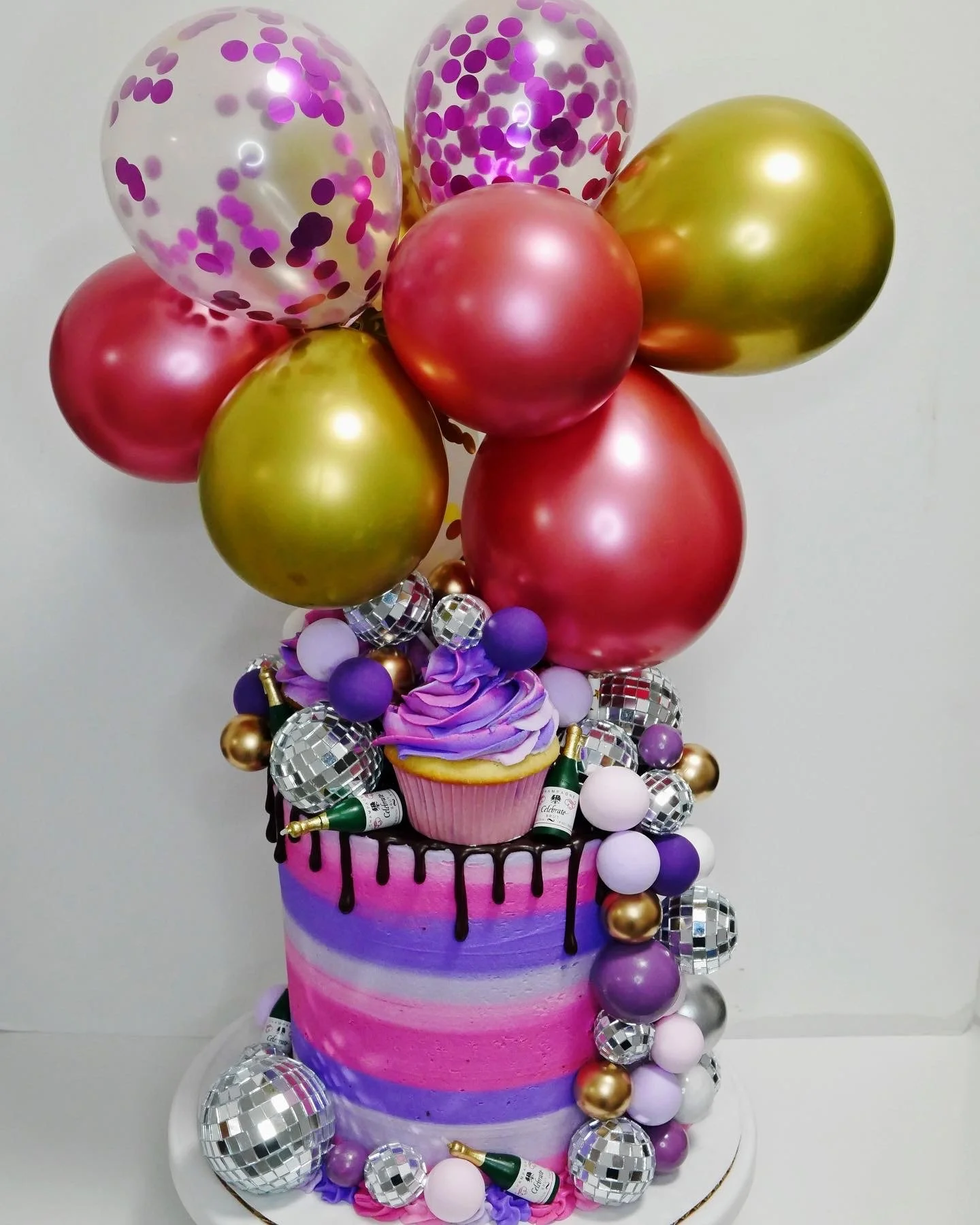 A cake decorated with purple, pink, and blue icing, topped with balloons in metallic gold, purple, pink, and clear with purple confetti, small disco balls, miniature champagne bottles, and a purple swirl cupcake with purple and pink icing.