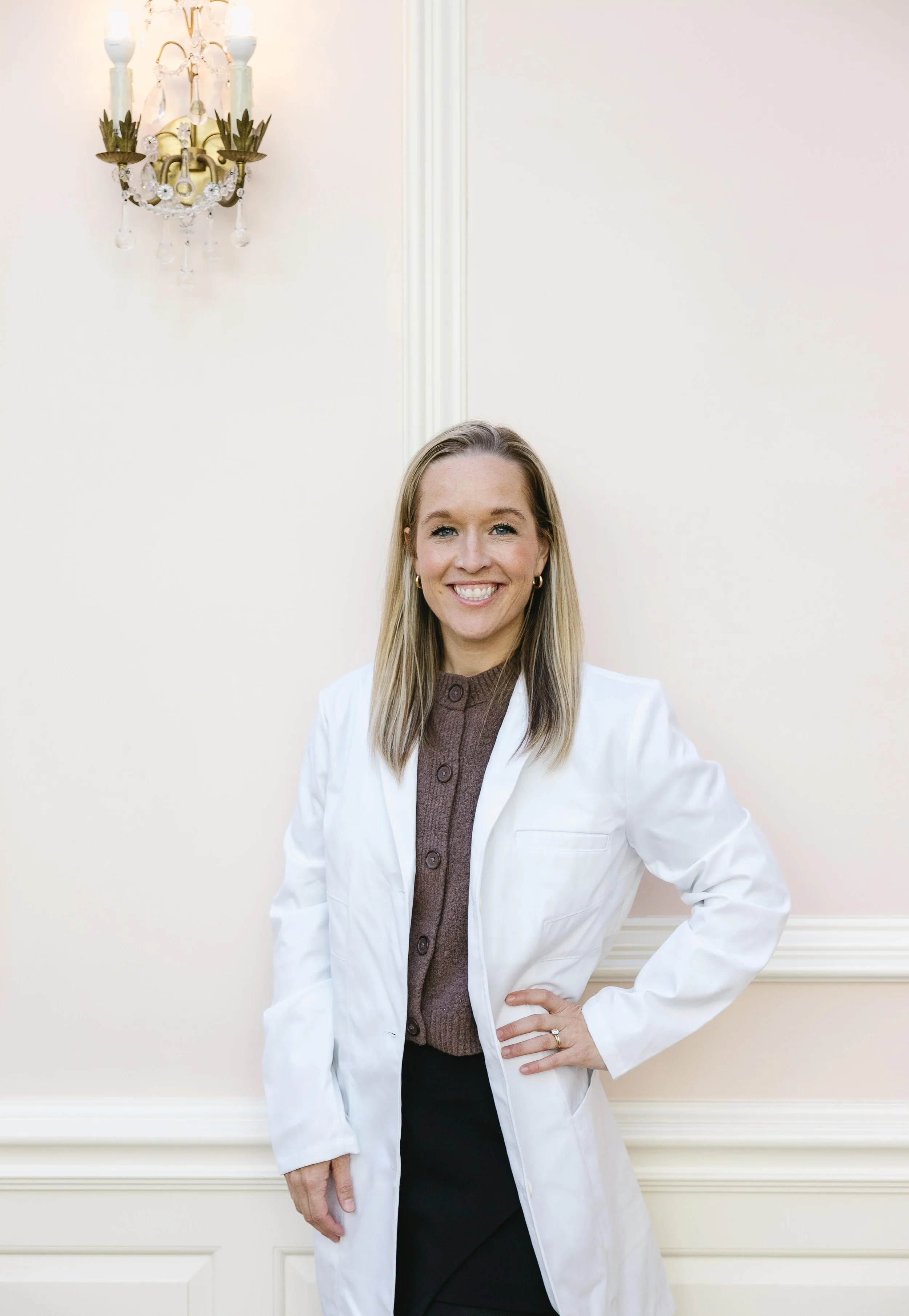 A woman in a white lab coat standing in front of a light pink wall, smiling and posing with one hand on her hip.