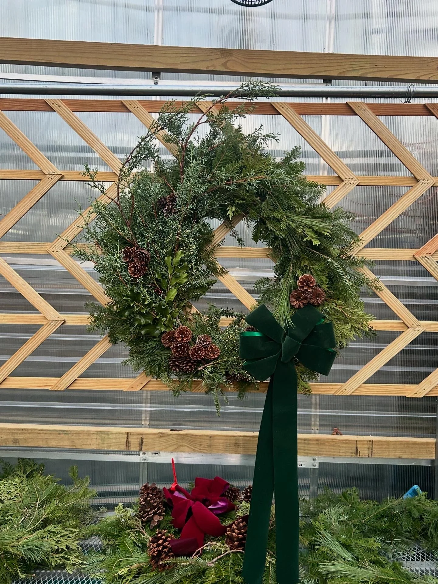 A holiday wreath decorated with pinecones, evergreen branches, and a green bow, displayed on a wooden lattice background.