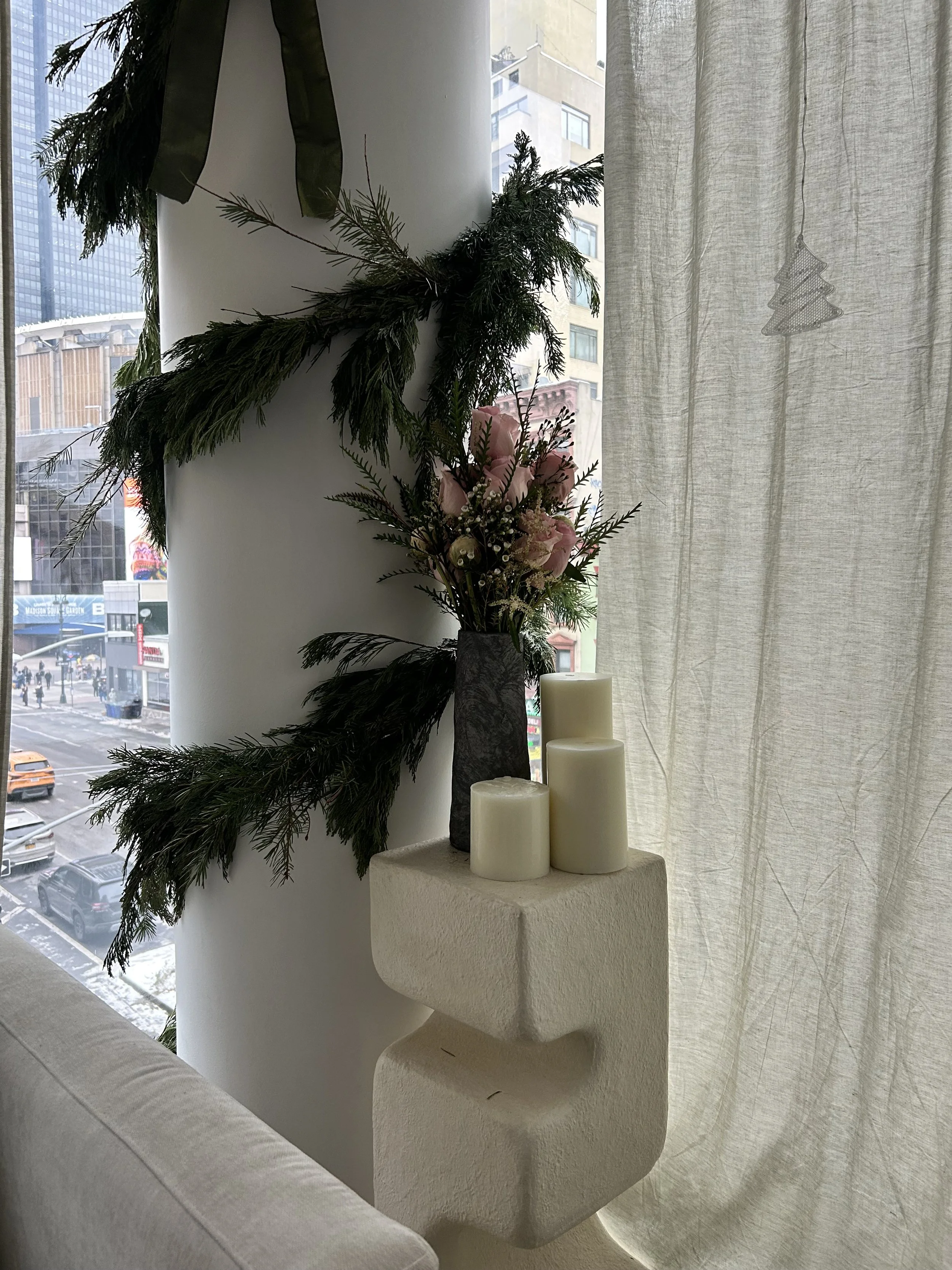 Interior decor with a flower arrangement and candles by a large window with sheer curtains, overlooking a city street.