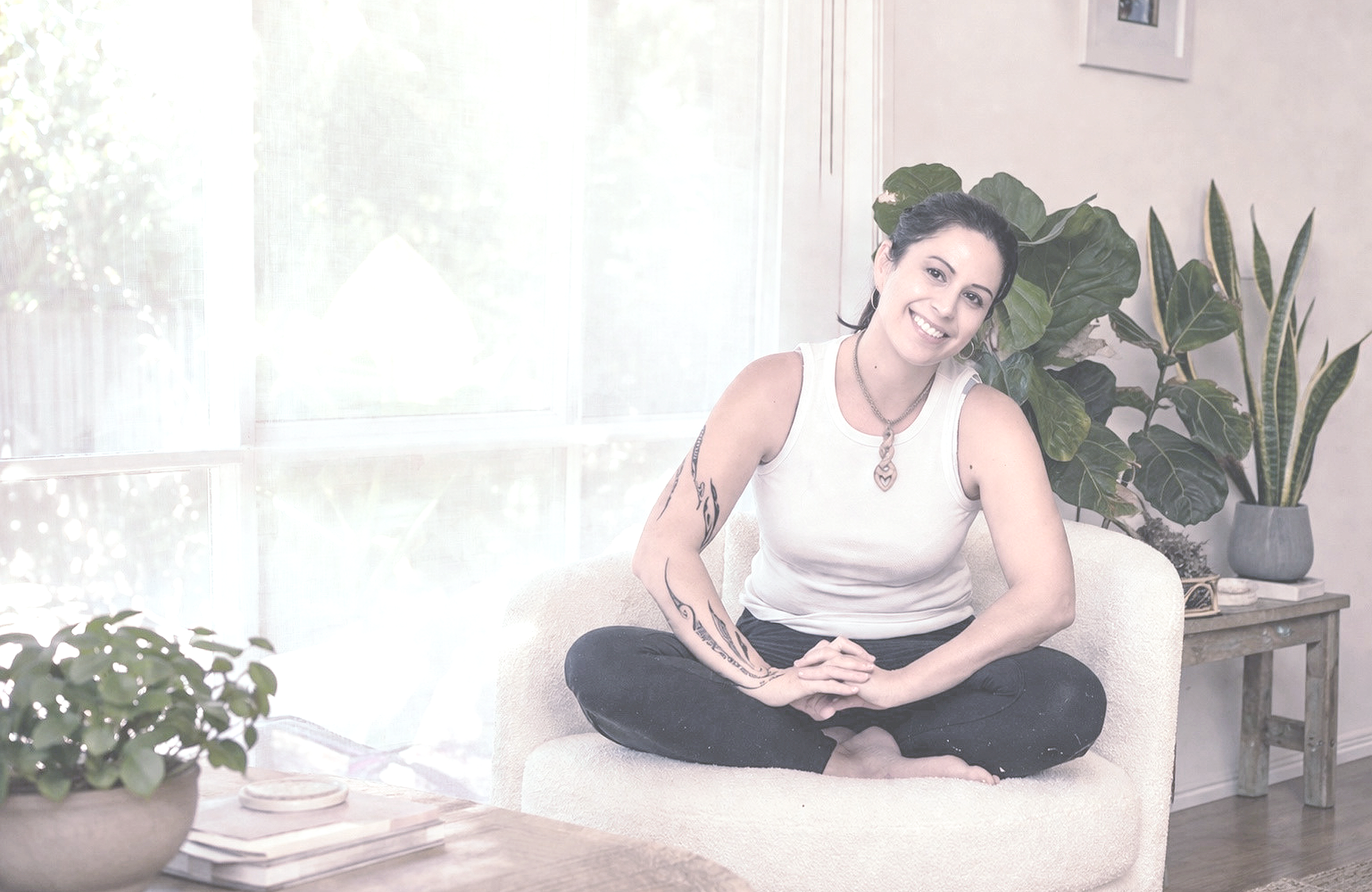 A woman sitting cross-legged on a cozy, cream-colored sofa in a sunlit room with large plants in the background. She has dark hair pulled back, tattoos on her arm, and is smiling at the camera.