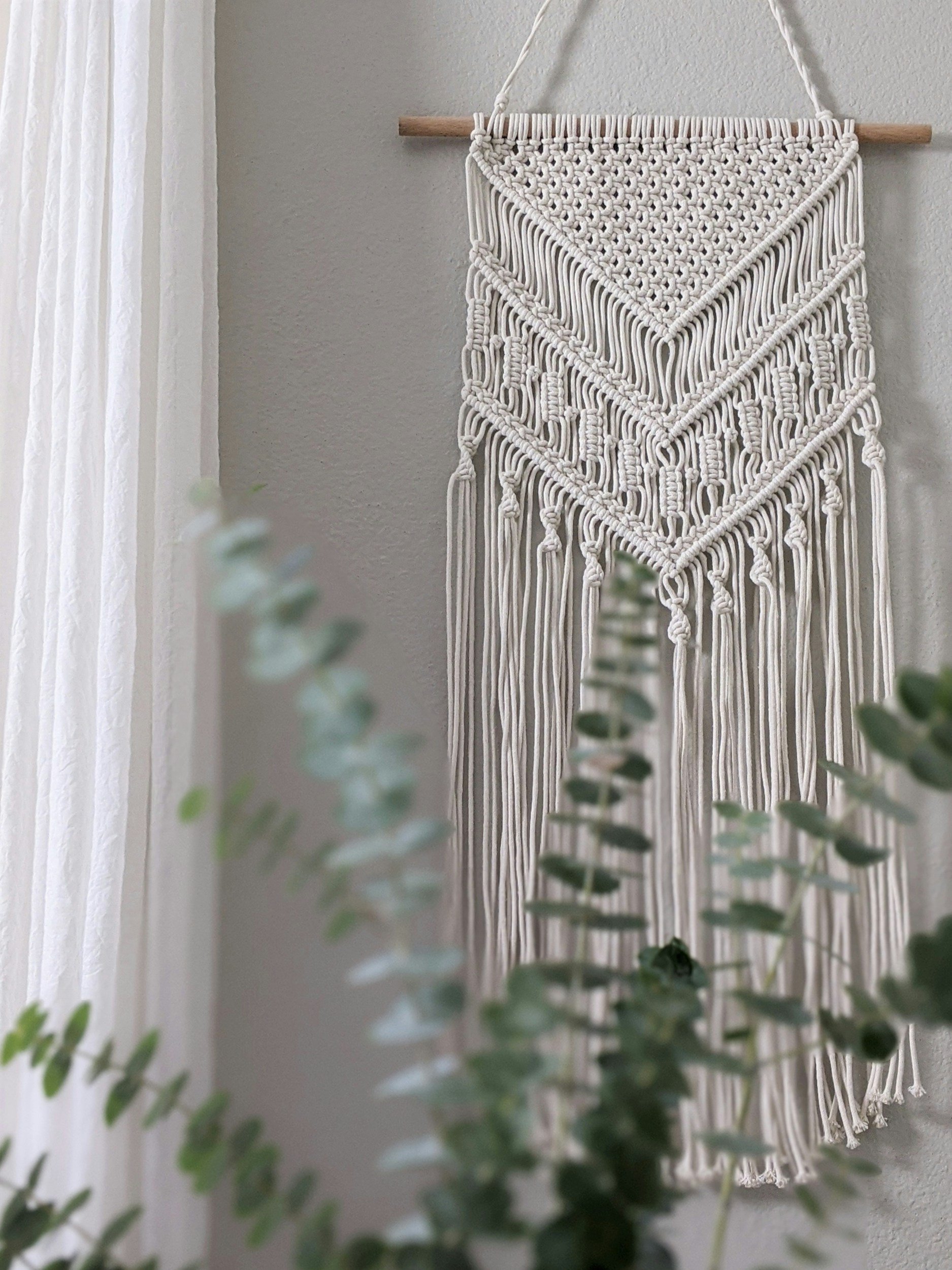 Decorative white macrame wall hanging with intricate knots and fringe, hanging on a wooden dowel against a gray wall with sheer white curtains and green plants in the foreground.