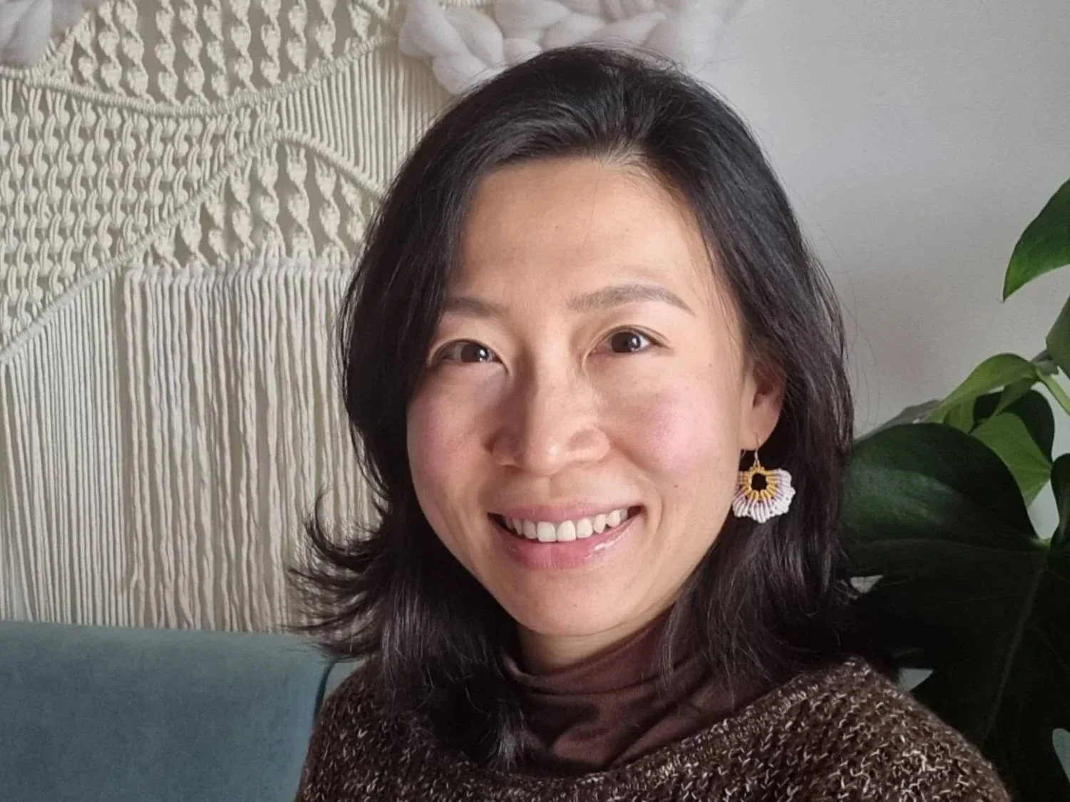 A woman smiling with dark, shoulder-length hair, wearing large hoop earrings and a brown patterned top, sitting indoors with a macrame wall hanging and a green plant in the background.