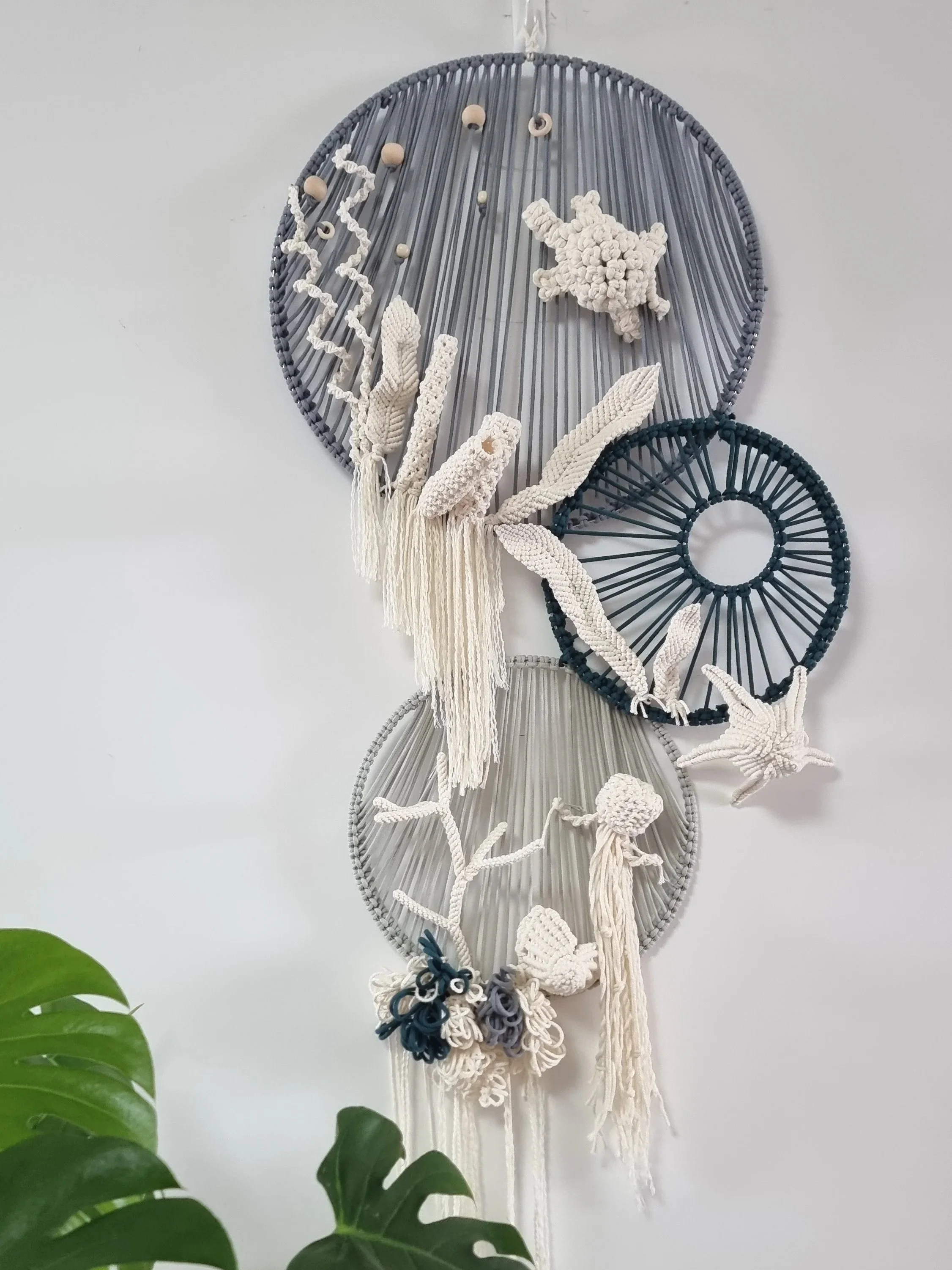 Decorative wall hanging made of large and small circular frames with woven, beaded, and macrame designs, featuring starfish, coral, and feather shapes, with green leaves visible in the foreground.