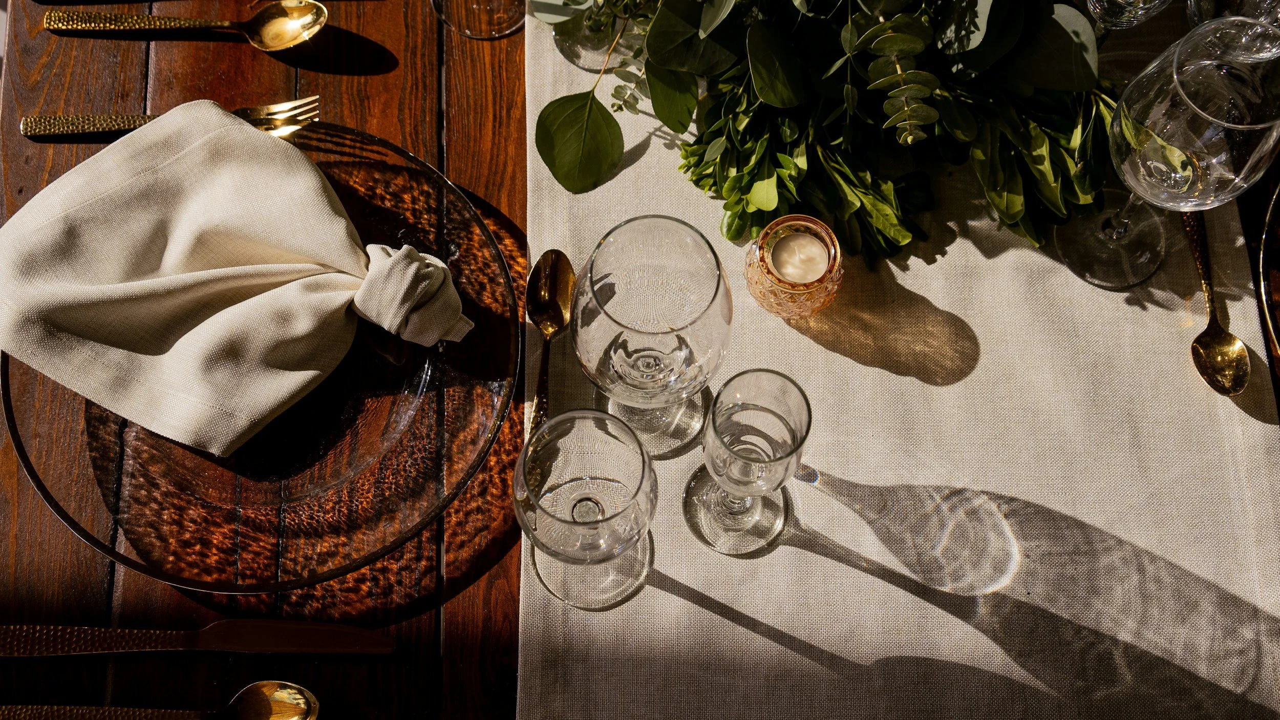 A table setting with a wooden table, a beige cloth napkin on a glass plate, gold utensils including a spoon, fork, and knife, several empty glasses, a small decorative candle holder, and a large green leafy plant at the center, with sunlight casting shadows.