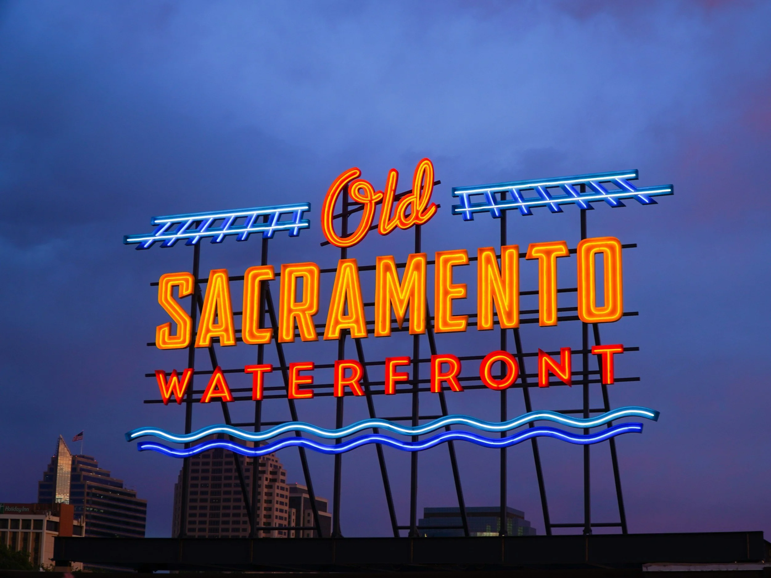 Cartel luminoso que dice 'Old Sacramento Waterfront' con fondo de ciudad y cielo nublado al atardecer.