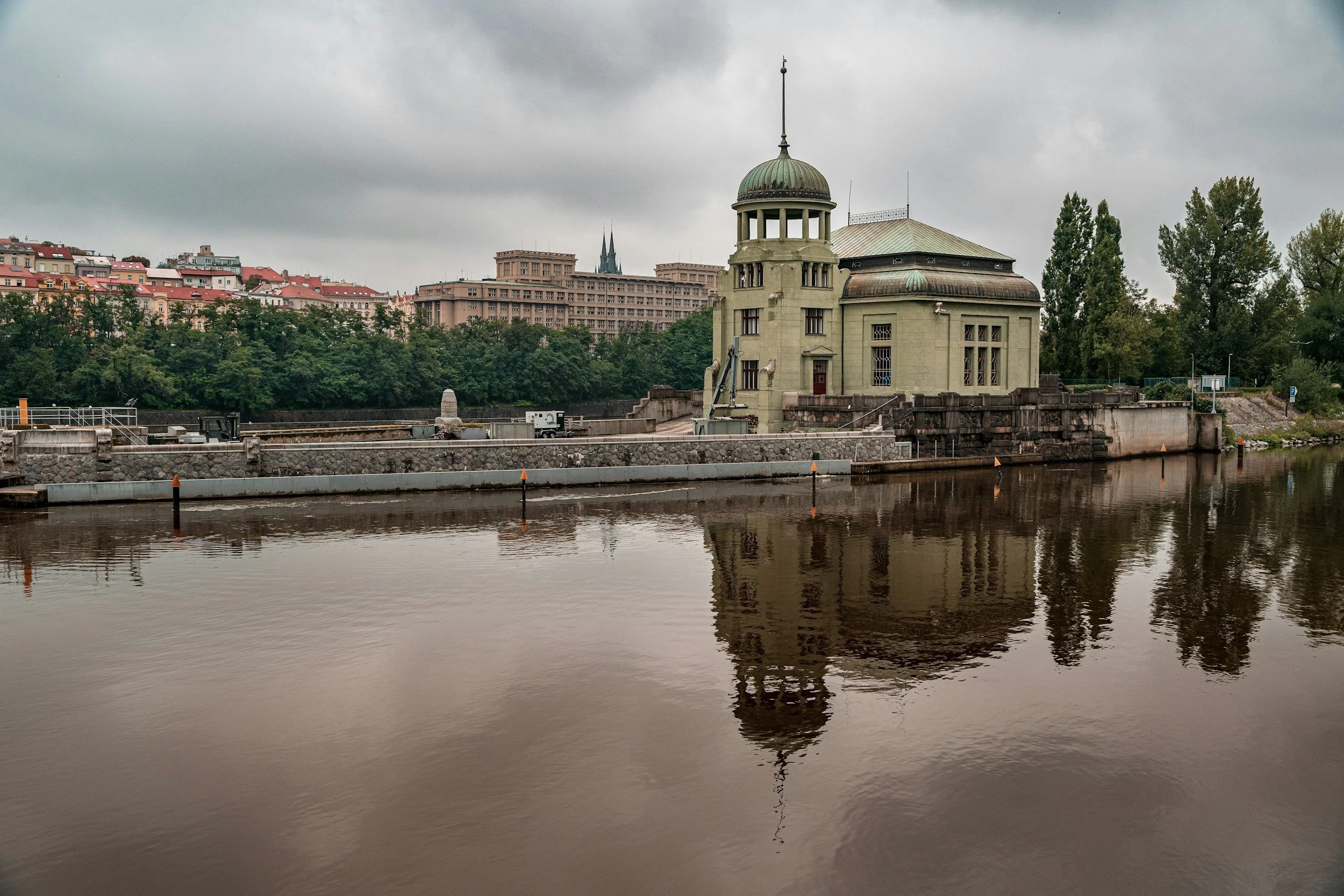 Edificio histórico junto al río, con árboles y edificios modernos al fondo en un día nublado.