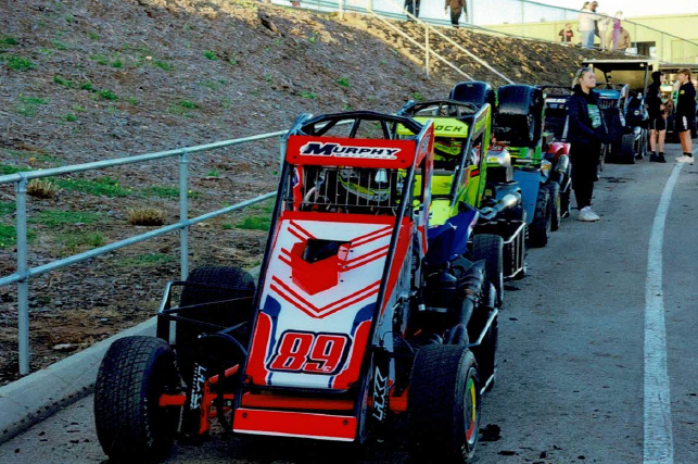 A racing go-kart with the number 89 and the name Murphy on the front, parked in a line with other go-karts and people standing nearby.