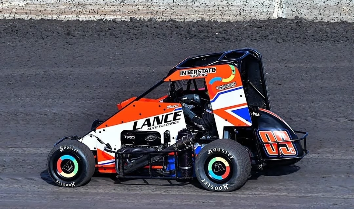 A sprint car race car on a dirt track, orange and black with the number 00 on the side, sponsor logos including Interstate Roofing and Lance Auto Electrics.
