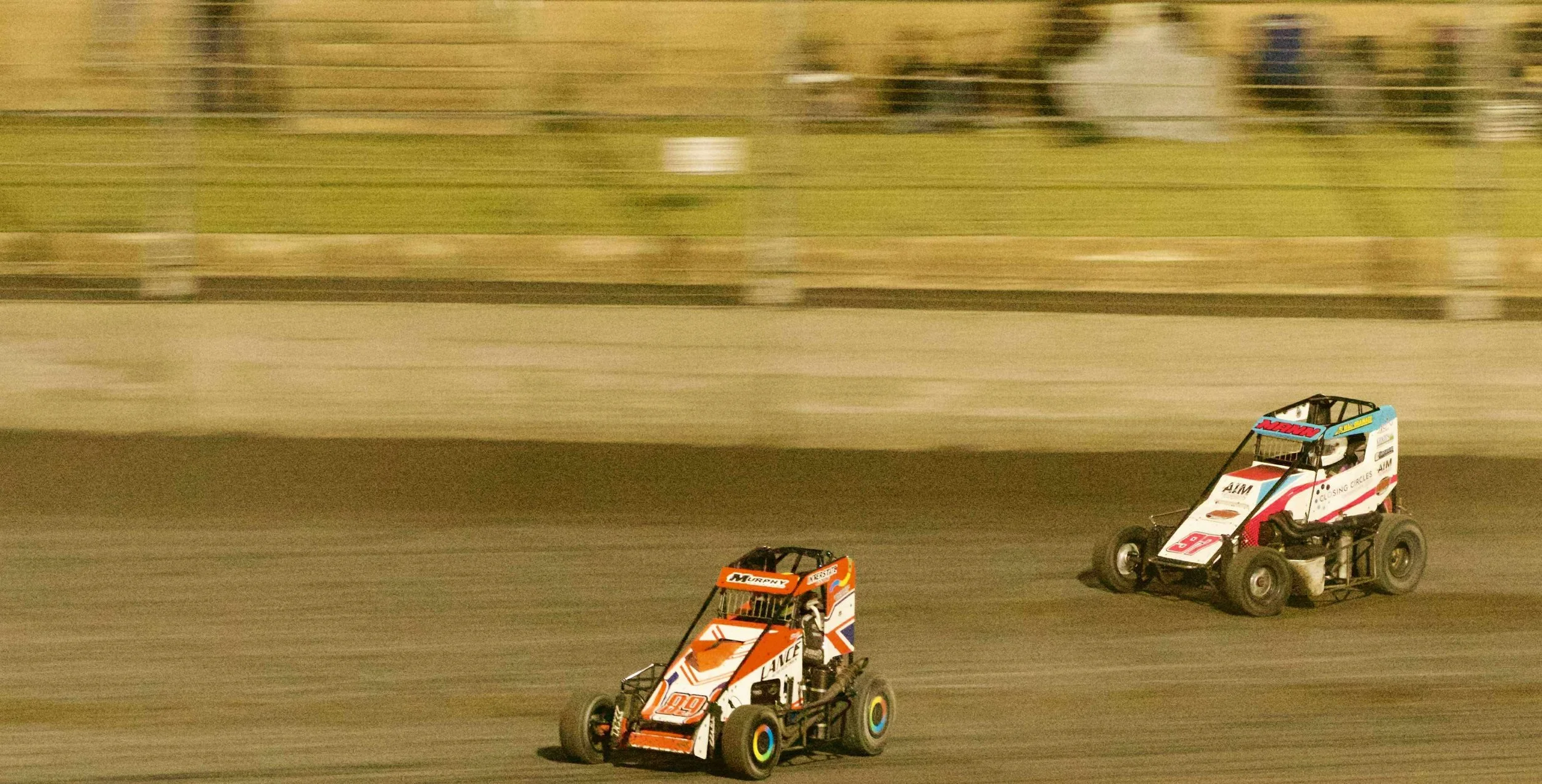 Two sprint cars racing on a dirt track at night.
