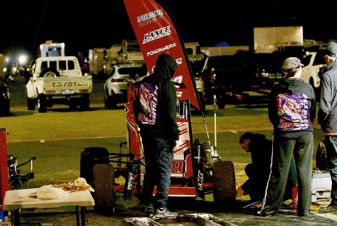 Group of people working on a race car at night, with tents, vehicles, and a food table in the background.