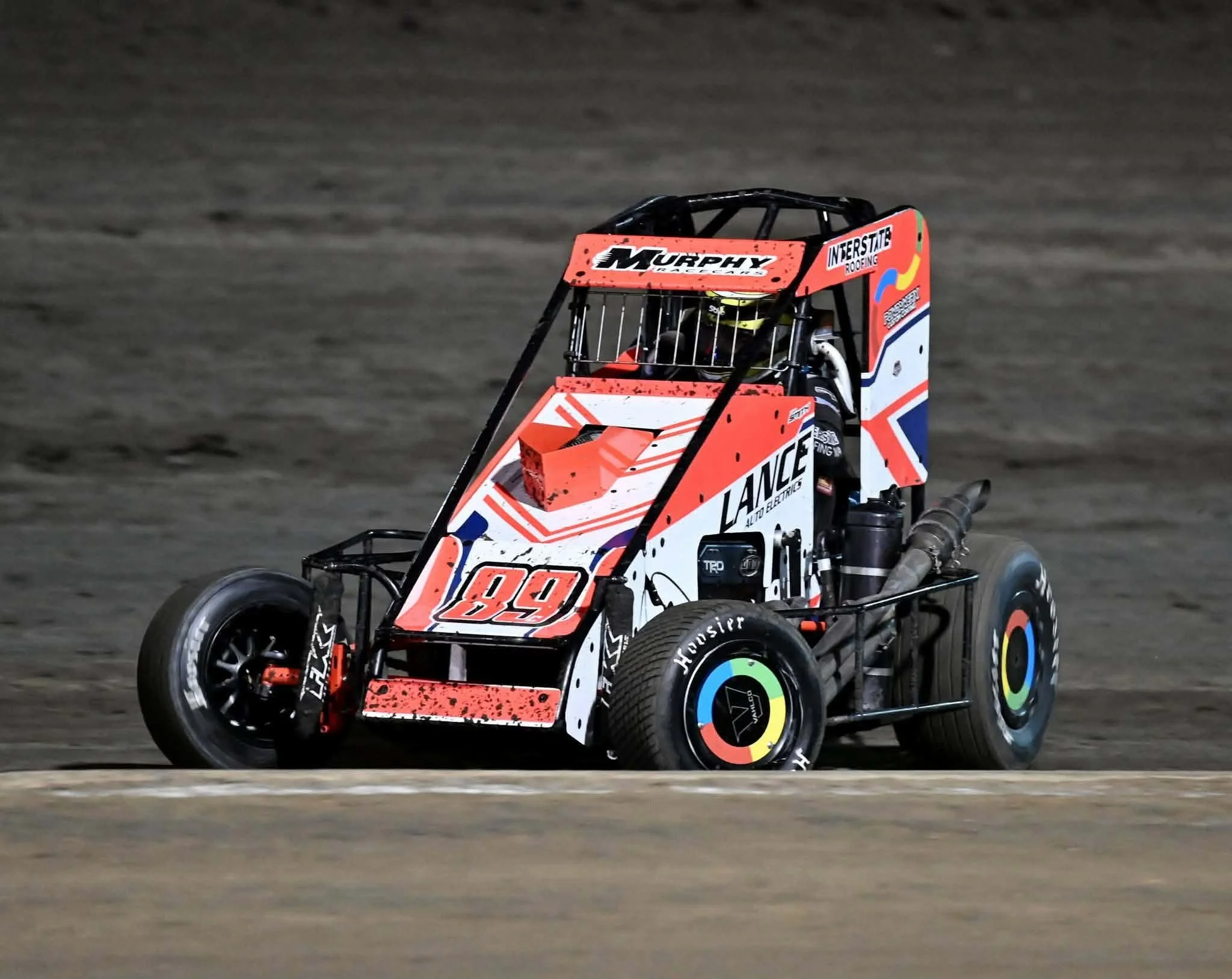 A sprint car racing on a dirt track at night, featuring a red, white, and blue color scheme, with the number 89 on the front and side, and various sponsor logos.