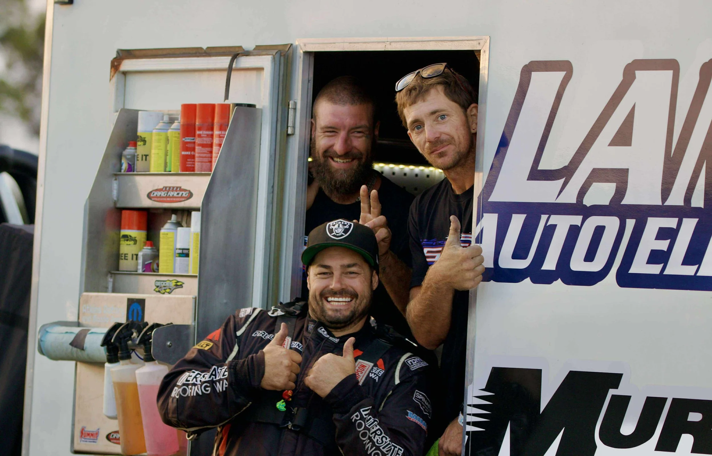 Three men, one in racing gear, smiling and giving thumbs up, standing in a promotional roadside auto show truck, with tools and products visible.