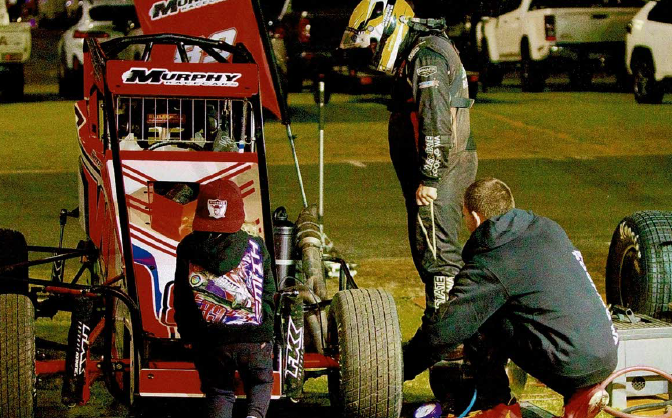 A group of people working on a red and black sprint car with the brand Murphy displayed on the wing. The scene takes place on a race track at night, with parked vehicles and a grassy area in the background.