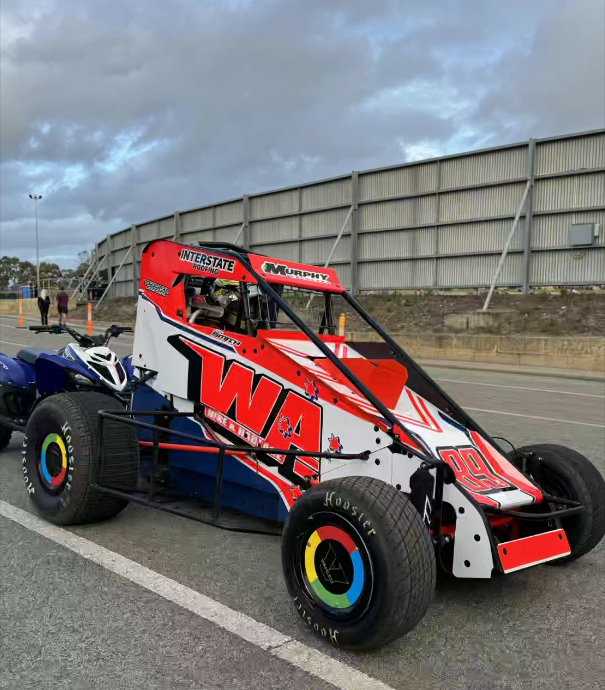 A red, white, and black race car, number 99, parked on a racetrack, with a winged structure and cloudy sky in the background.